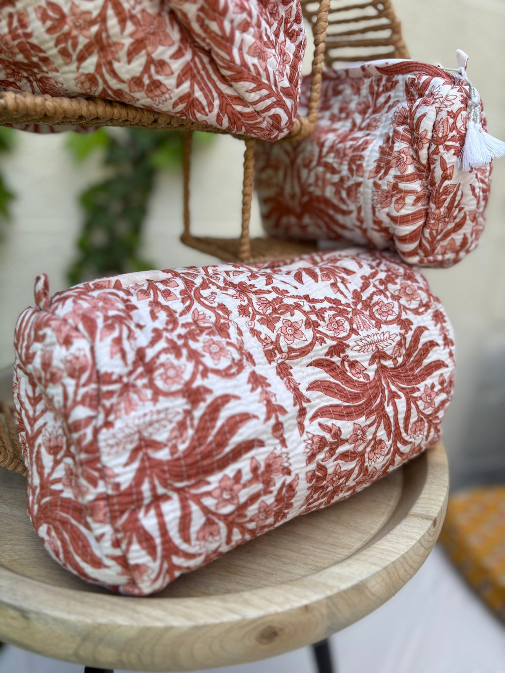 Un ensemble de trois trousses de toilette cylindriques en coton matelassé (technique Kantha) est présenté sur une étagère circulaire en osier tressé. Le tissu blanc est orné d'un imprimé blockprint monochrome dans des tons terre cuite et brique, représentant des motifs floraux et des feuillages denses. La texture est marquée par des surpiqûres horizontales régulières. La scène, capturée en extérieur avec un arrière-plan de lierre vert sur un mur clair, met en valeur le caractère artisanal et naturel 