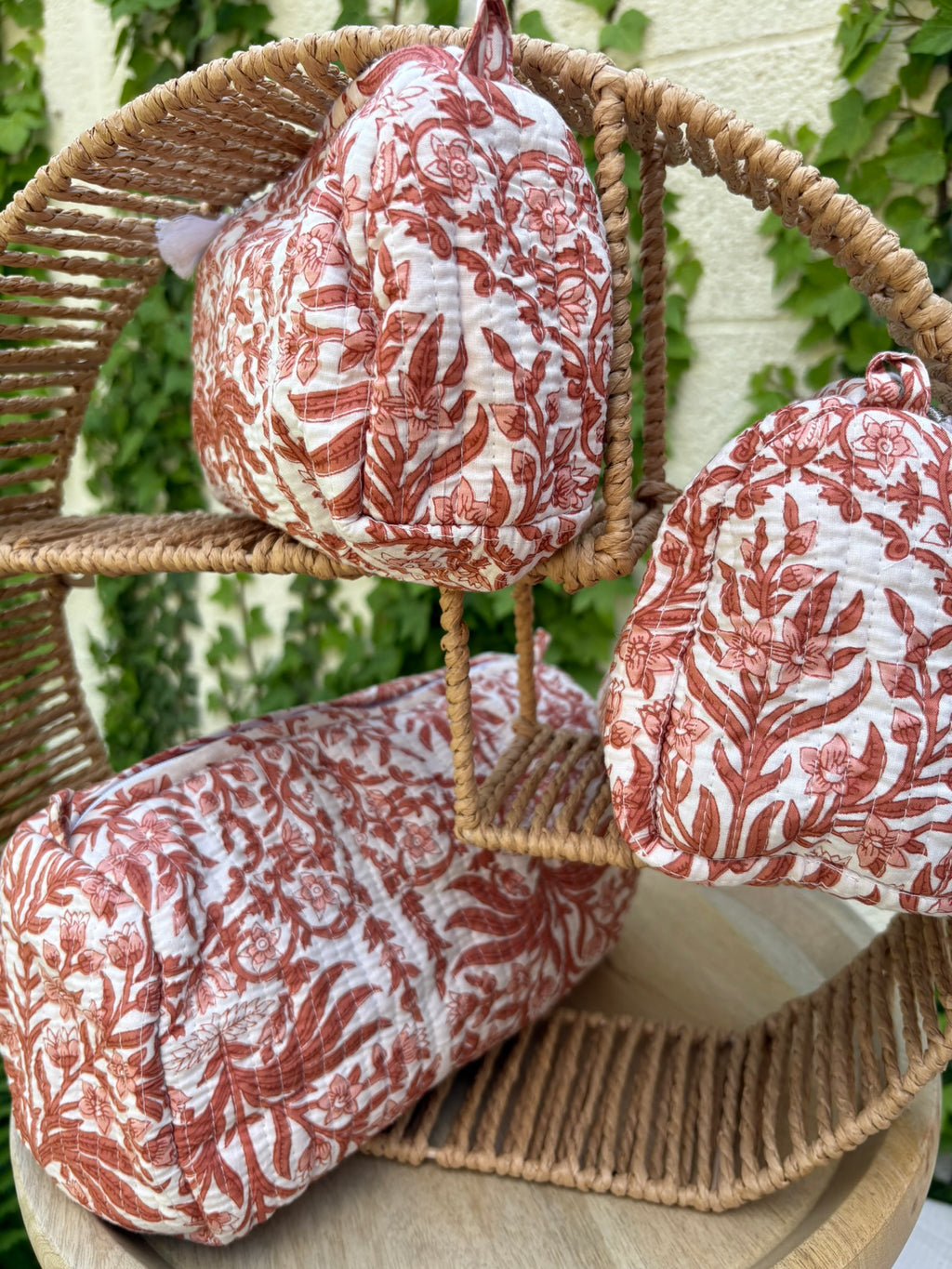 Un ensemble de trois trousses de toilette cylindriques en coton matelassé (technique Kantha) est présenté sur une étagère circulaire en osier tressé. Le tissu blanc est orné d'un imprimé blockprint monochrome dans des tons terre cuite et brique, représentant des motifs floraux et des feuillages denses. La texture est marquée par des surpiqûres horizontales régulières. La scène, capturée en extérieur avec un arrière-plan de lierre vert sur un mur clair, met en valeur le caractère artisanal et naturel 