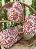 Un ensemble de trois trousses de toilette cylindriques en coton matelassé (technique Kantha) est présenté sur une étagère circulaire en osier tressé. Le tissu blanc est orné d'un imprimé blockprint monochrome dans des tons terre cuite et brique, représentant des motifs floraux et des feuillages denses. La texture est marquée par des surpiqûres horizontales régulières. La scène, capturée en extérieur avec un arrière-plan de lierre vert sur un mur clair, met en valeur le caractère artisanal et naturel 