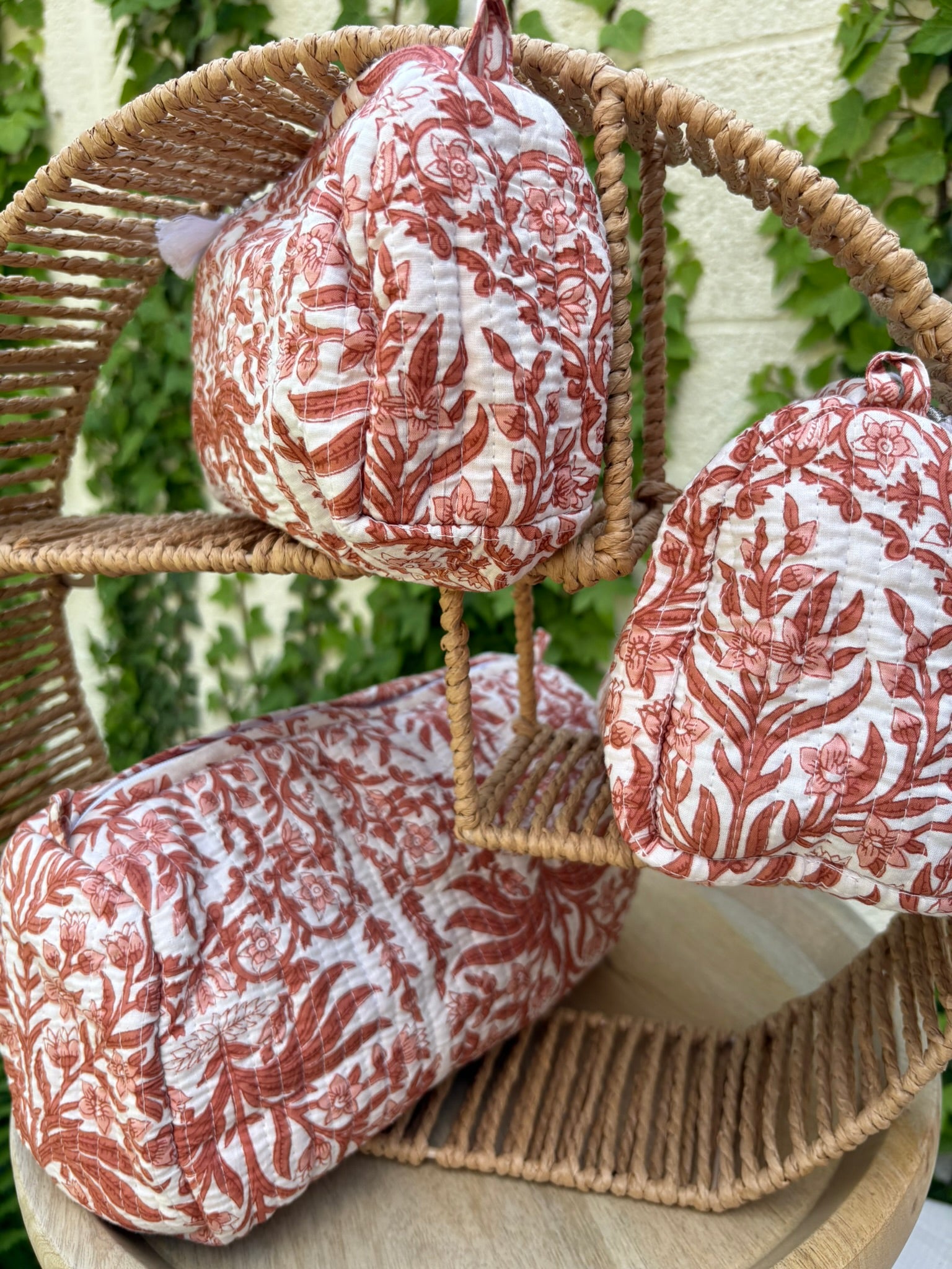 Un ensemble de trois trousses de toilette cylindriques en coton matelassé (technique Kantha) est présenté sur une étagère circulaire en osier tressé. Le tissu blanc est orné d'un imprimé blockprint monochrome dans des tons terre cuite et brique, représentant des motifs floraux et des feuillages denses. La texture est marquée par des surpiqûres horizontales régulières. La scène, capturée en extérieur avec un arrière-plan de lierre vert sur un mur clair, met en valeur le caractère artisanal et naturel 