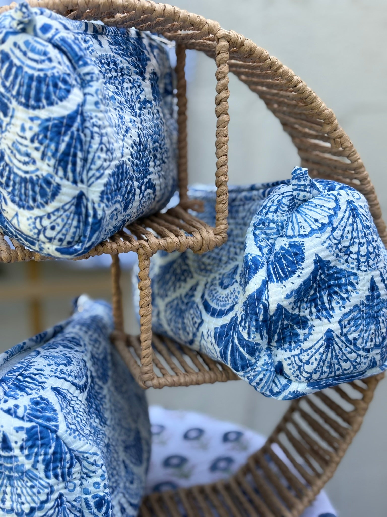 Trousses de toilette matelassées en coton blanc avec motifs coquillage bleu indigo imprimés au blockprint artisanal, présentées sur une étagère ronde en rotin tressé.