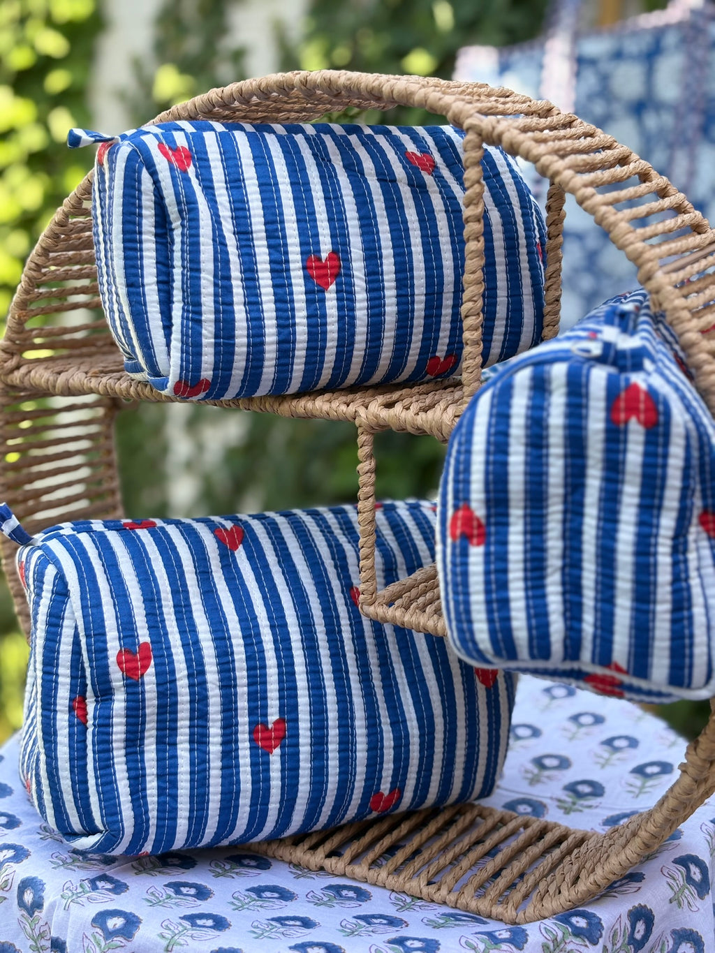 Trois trousses de toilette en coton matelassé bleu et blanc à rayures, imprimées de petits cœurs rouges en blockprint artisanal, présentées sur une étagère en rotin tressé.