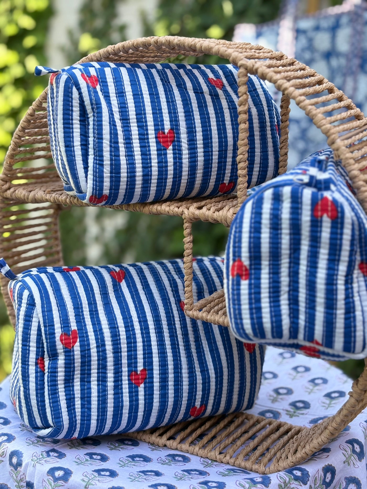Trois trousses de toilette en coton matelassé bleu et blanc à rayures, imprimées de petits cœurs rouges en blockprint artisanal, présentées sur une étagère en rotin tressé.