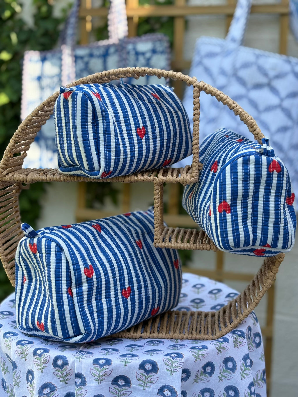 Trois trousses de toilette en coton matelassé bleu et blanc à rayures, imprimées de petits cœurs rouges en blockprint artisanal, présentées sur une étagère en rotin tressé.