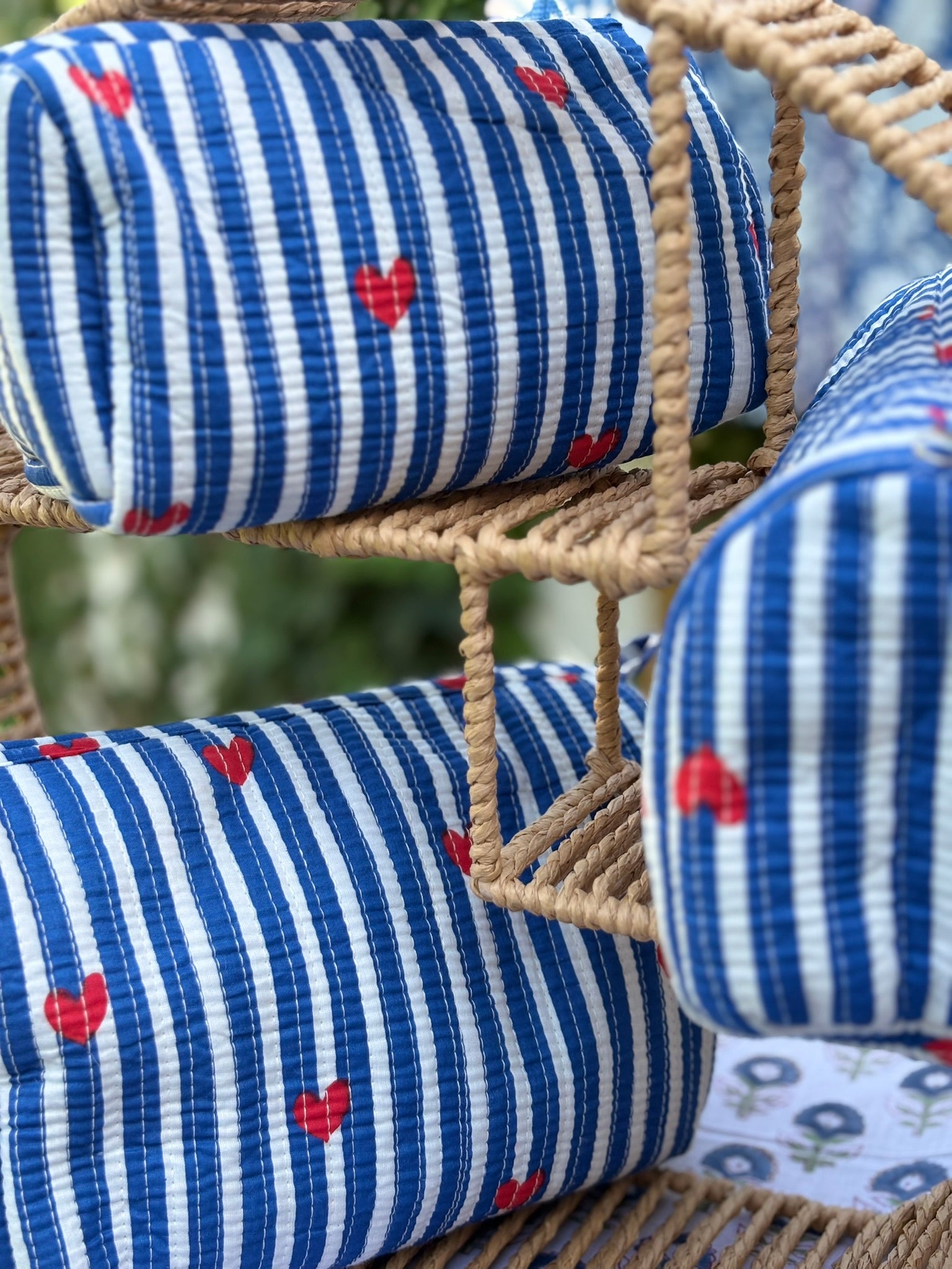Trois trousses de toilette en coton matelassé bleu et blanc à rayures, imprimées de petits cœurs rouges en blockprint artisanal, présentées sur une étagère en rotin tressé.