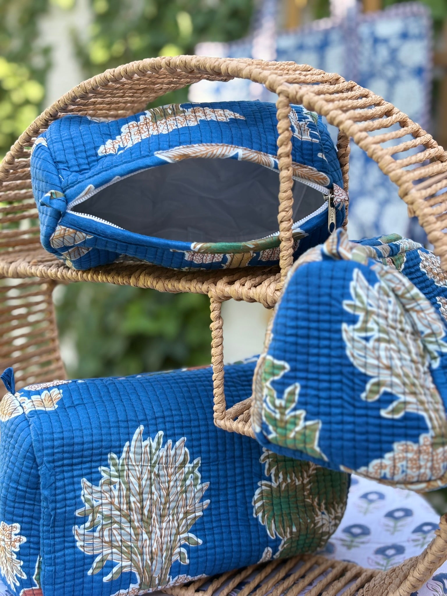 Trois trousses de toilette en coton matelassé bleu canard avec motifs d'arbres et feuillages végétaux ocre et vert, imprimées en blockprint artisanal sur une étagère en rotin