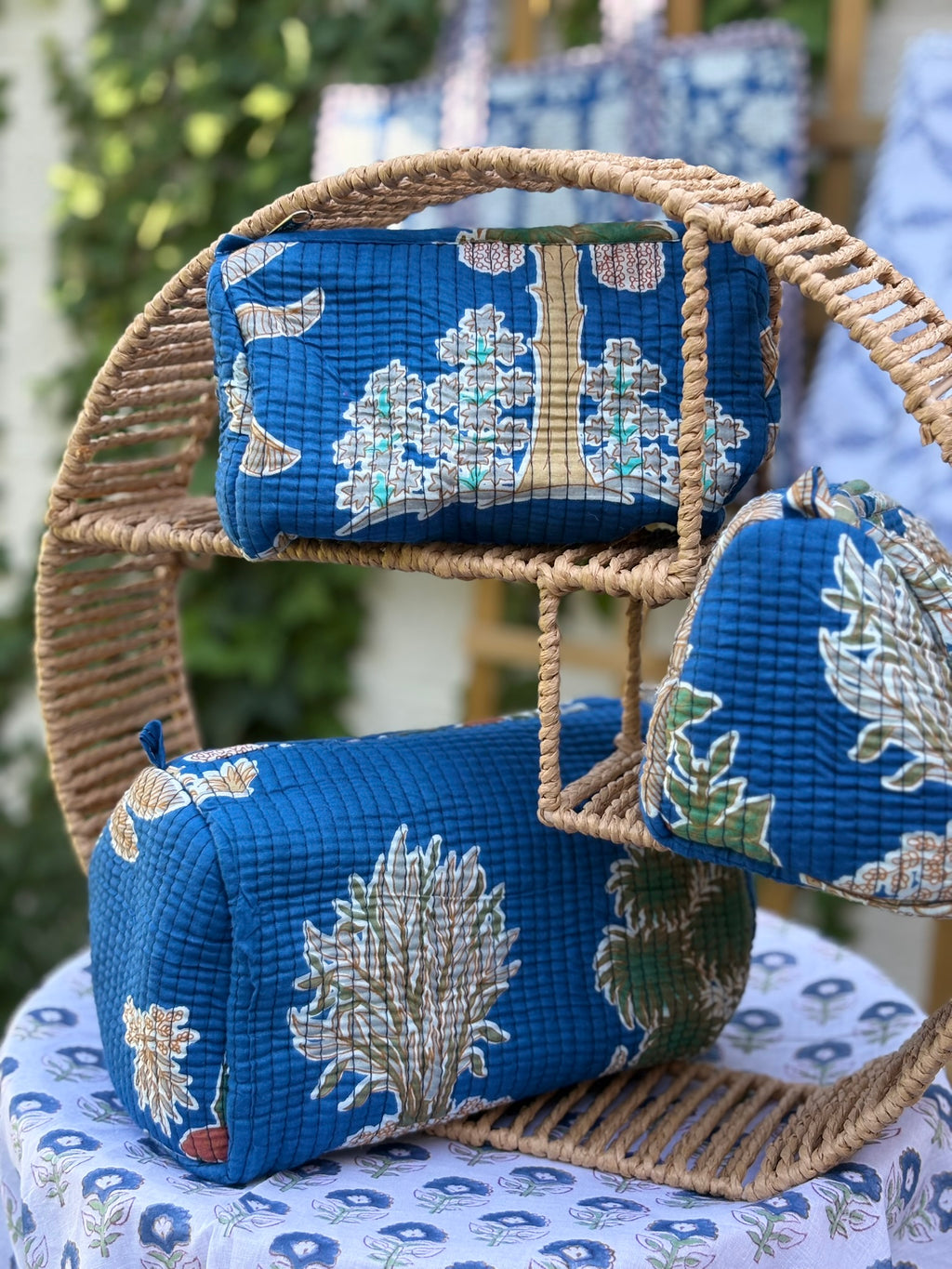 Trois trousses de toilette en coton matelassé bleu canard avec motifs d'arbres et feuillages végétaux ocre et vert, imprimées en blockprint artisanal sur une étagère en rotin