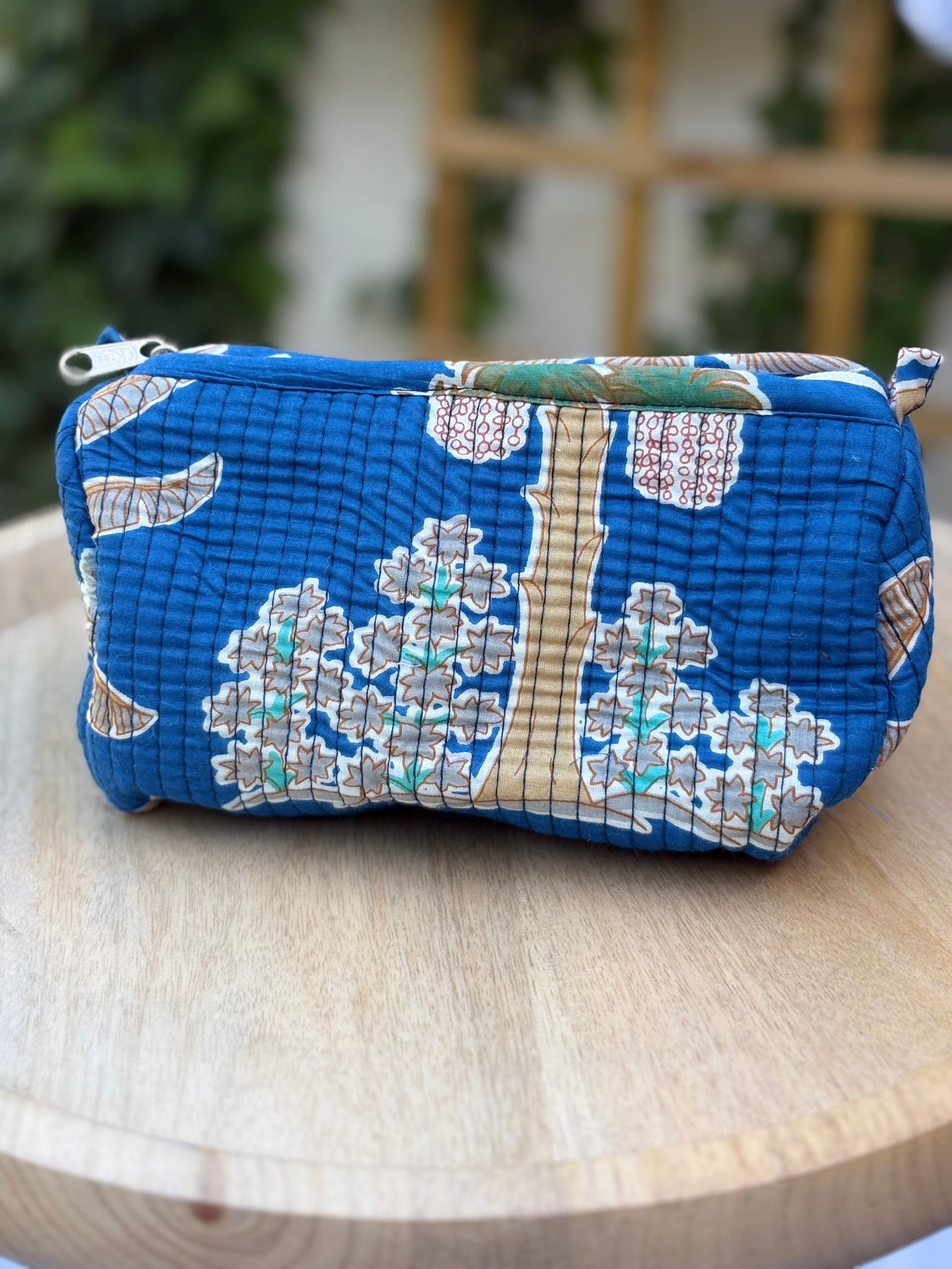 Une trousse de toilette en coton matelassé bleu avec motifs d'arbres et feuillages végétaux ocre et vert, imprimée en blockprint artisanal sur une table ronde