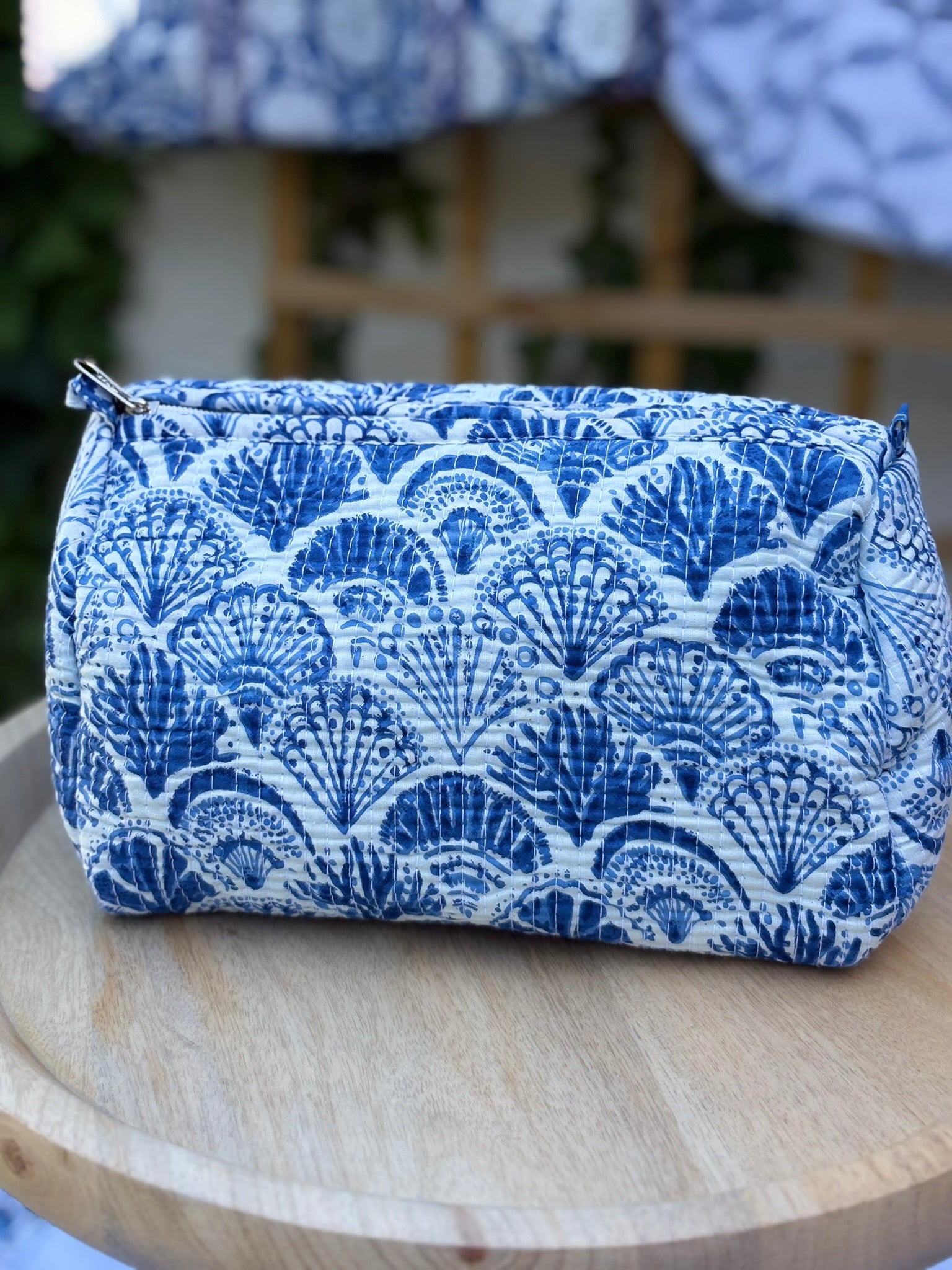 Trousse de toilette matelassée en coton blanc avec motifs coquillage bleu indigo imprimés au blockprint artisanal, présentée sur une étagère ronde en rotin tressé.