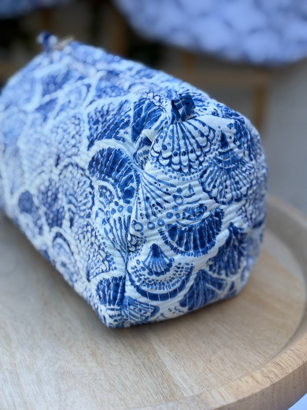 Trousse de toilette matelassée en coton blanc avec motifs coquillage bleu indigo imprimés au blockprint artisanal, présentée sur une étagère ronde en rotin tressé.
