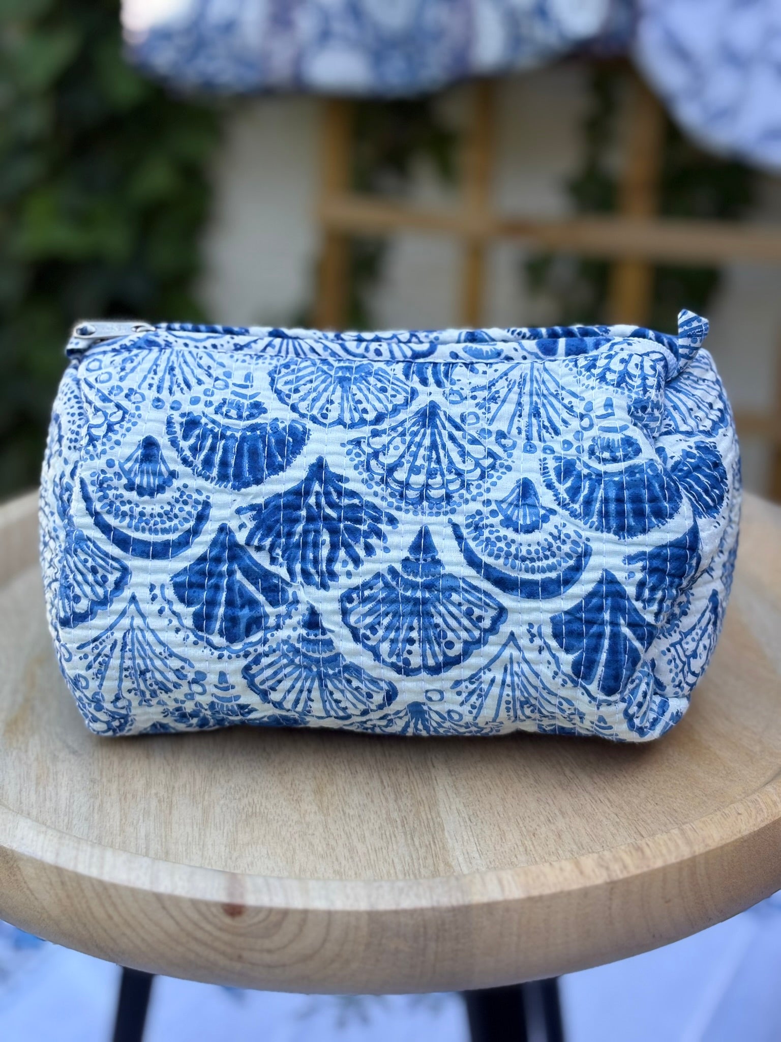 Trousse de toilette matelassée en coton blanc avec motifs coquillage bleu indigo imprimés au blockprint artisanal, présentée sur une étagère ronde en rotin tressé.