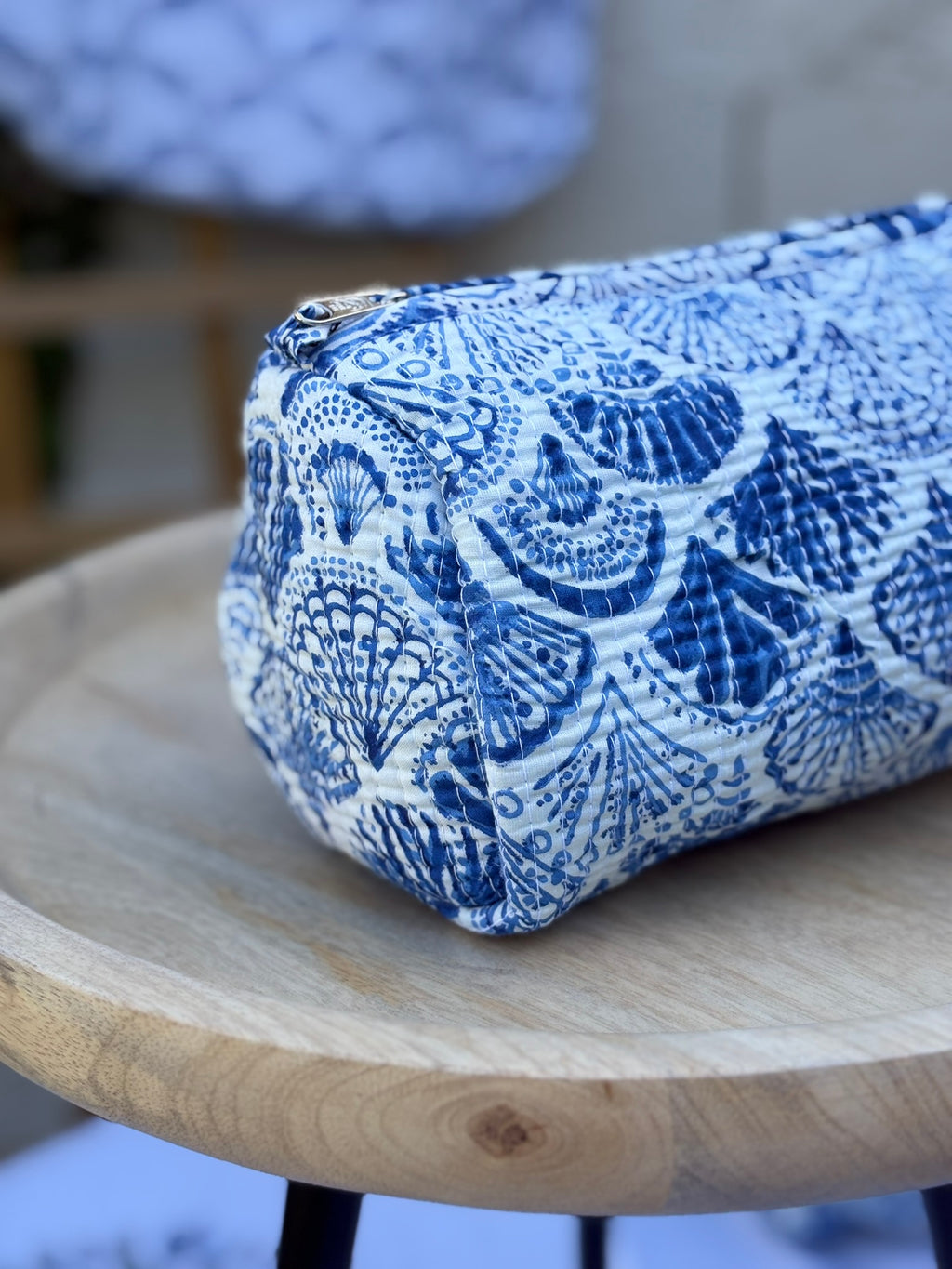 Trousse de toilette matelassée en coton blanc avec motifs coquillage bleu indigo imprimés au blockprint artisanal, présentée sur une étagère ronde en rotin tressé.