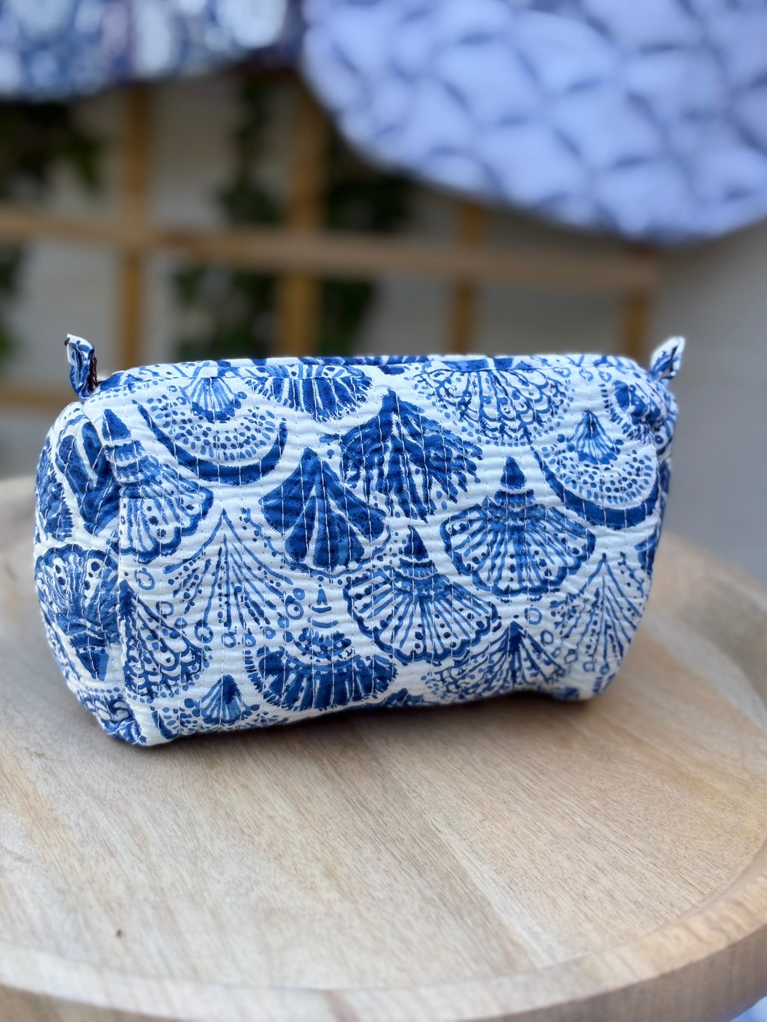 Trousse de toilette matelassée en coton blanc avec motifs coquillage bleu indigo imprimés au blockprint artisanal, présentée sur une étagère ronde en rotin tressé.