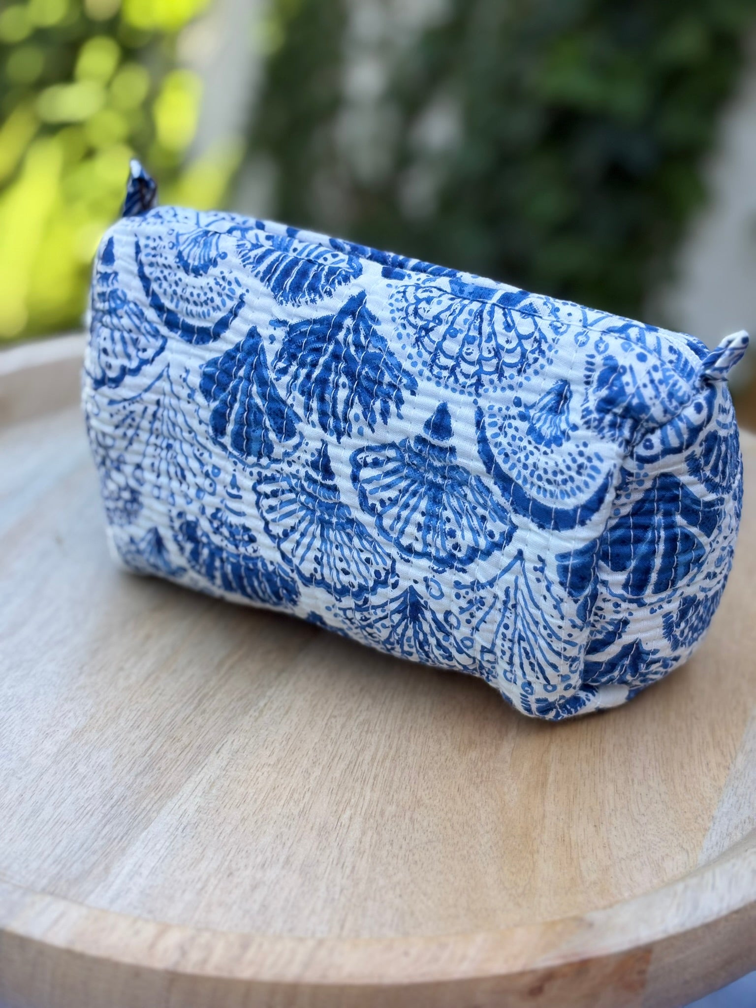 Trousse de toilette matelassée en coton blanc avec motifs coquillage bleu indigo imprimés au blockprint artisanal, présentée sur une étagère ronde en rotin tressé.