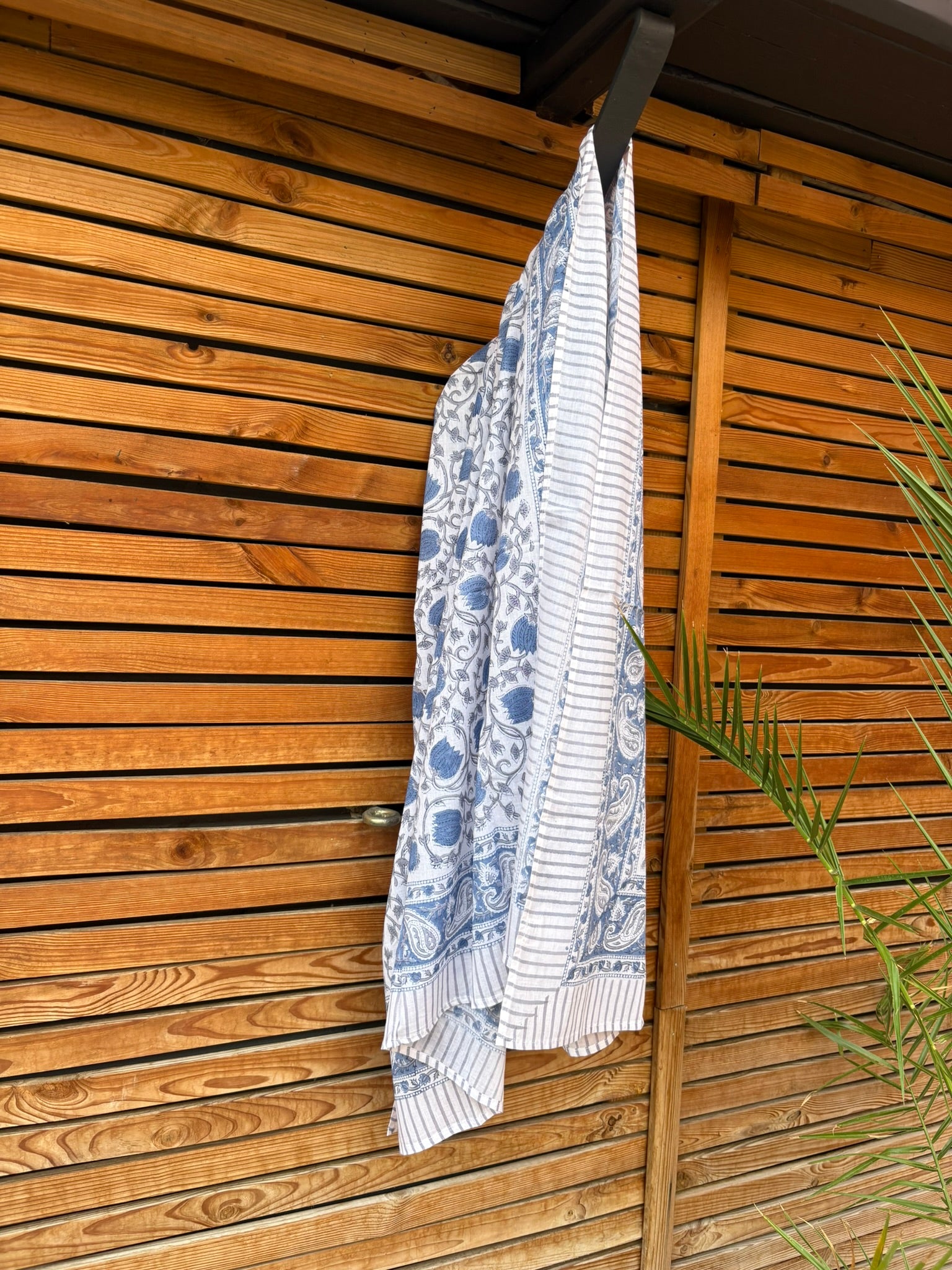 Paréo en coton léger avec impression artisanale indienne de type block print. Il présente un motif floral et végétal détaillé aux teintes bleu ardoise et gris sur un fond blanc, structuré par de larges bordures ornées et des finitions à fines rayures. Le tissu est suspendu naturellement devant un mur à claire-voie en bois clair et un feuillage vert
