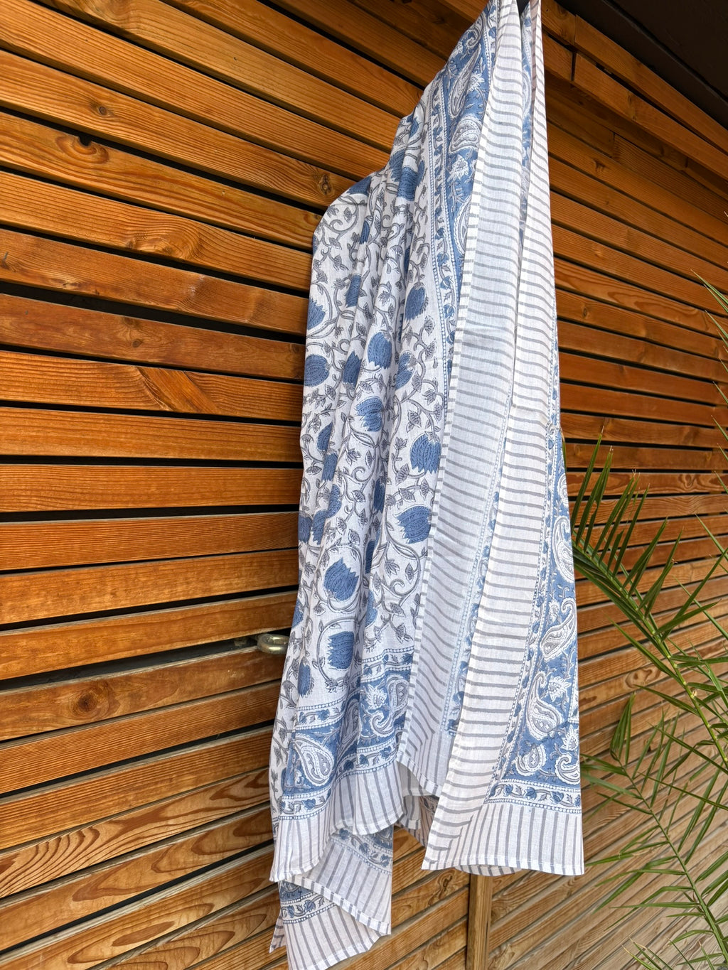 Paréo en coton léger avec impression artisanale indienne de type block print. Il présente un motif floral et végétal détaillé aux teintes bleu ardoise et gris sur un fond blanc, structuré par de larges bordures ornées et des finitions à fines rayures. Le tissu est suspendu naturellement devant un mur à claire-voie en bois clair et un feuillage vert