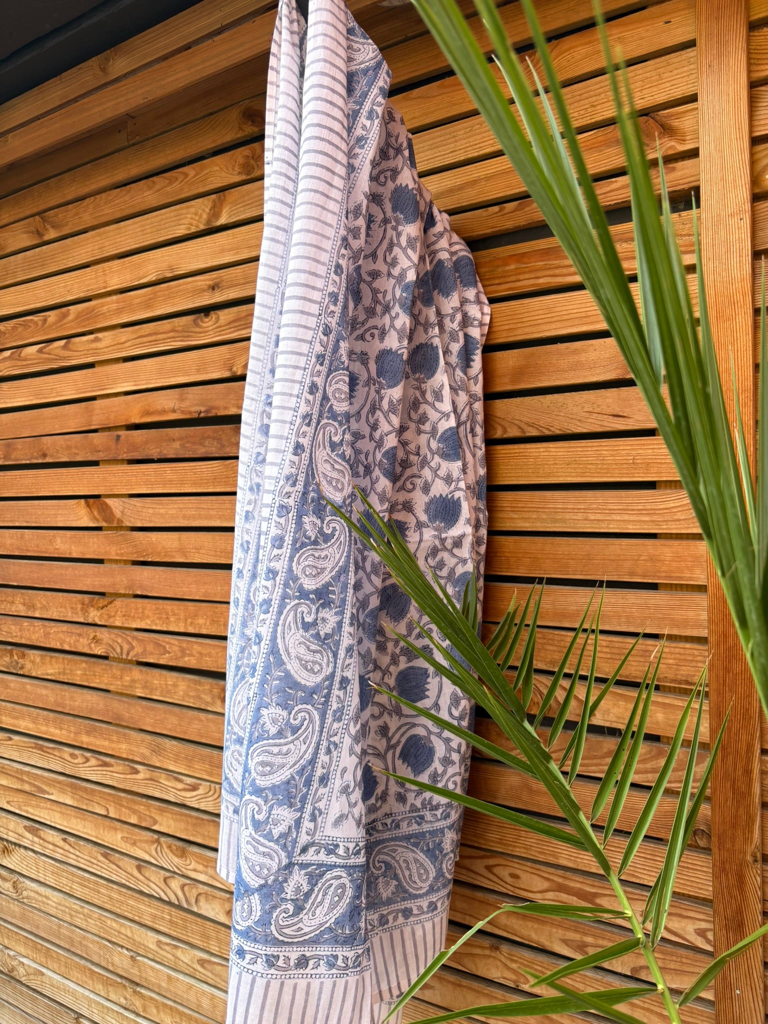 Paréo en coton léger avec impression artisanale indienne de type block print. Il présente un motif floral et végétal détaillé aux teintes bleu ardoise et gris sur un fond blanc, structuré par de larges bordures ornées et des finitions à fines rayures. Le tissu est suspendu naturellement devant un mur à claire-voie en bois clair et un feuillage vert