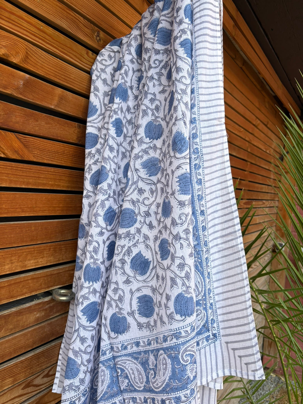 Paréo en coton léger avec impression artisanale indienne de type block print. Il présente un motif floral et végétal détaillé aux teintes bleu ardoise et gris sur un fond blanc, structuré par de larges bordures ornées et des finitions à fines rayures. Le tissu est suspendu naturellement devant un mur à claire-voie en bois clair et un feuillage vert