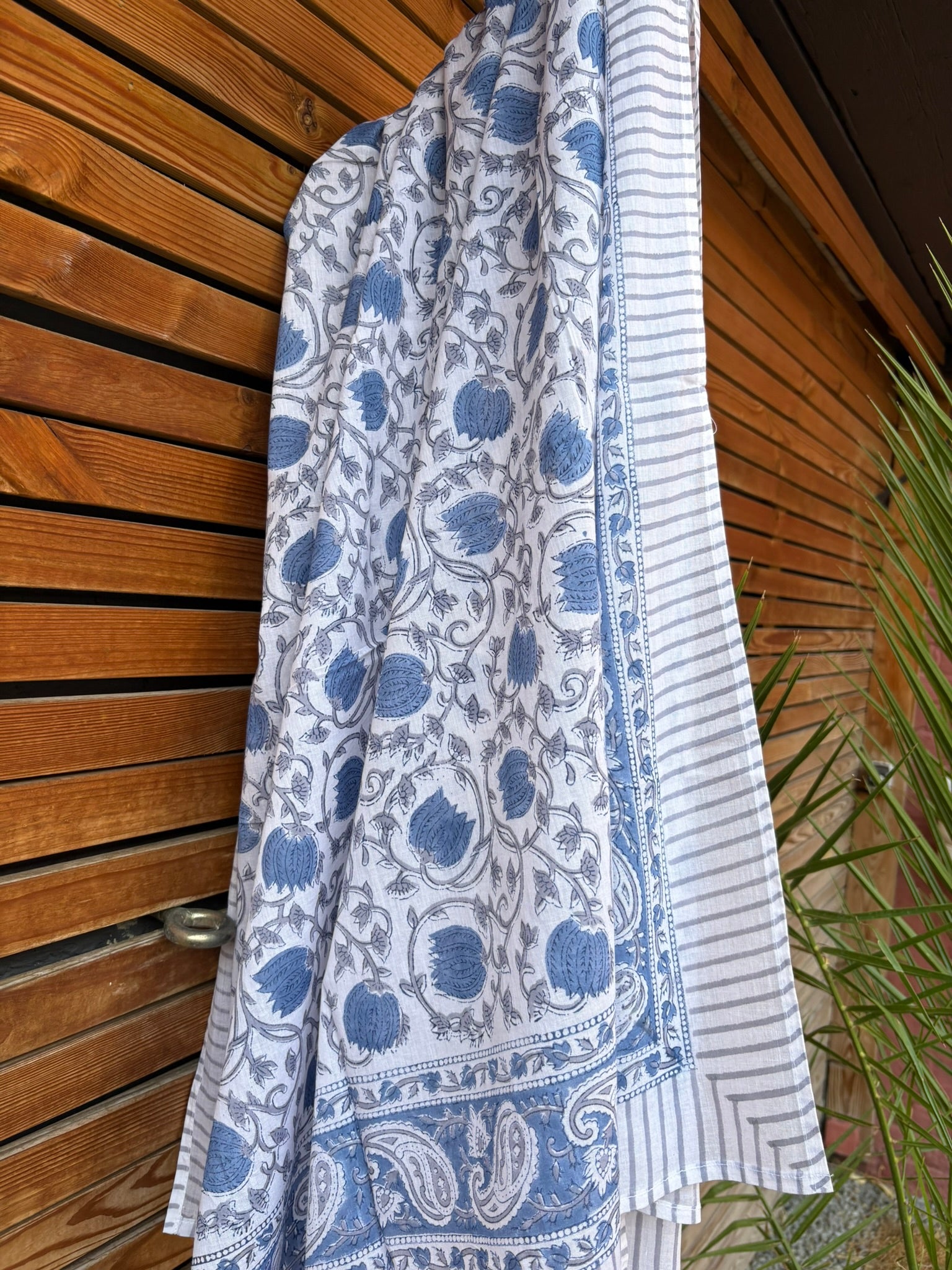 Paréo en coton léger avec impression artisanale indienne de type block print. Il présente un motif floral et végétal détaillé aux teintes bleu ardoise et gris sur un fond blanc, structuré par de larges bordures ornées et des finitions à fines rayures. Le tissu est suspendu naturellement devant un mur à claire-voie en bois clair et un feuillage vert