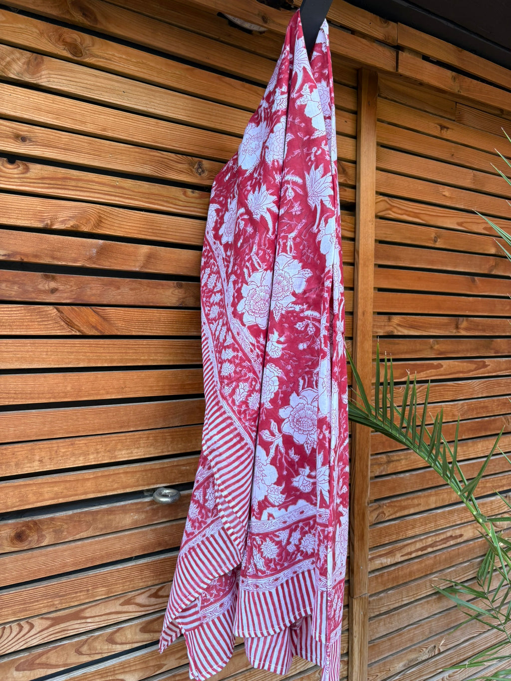 Paréo en coton léger avec impression artisanale indienne de type block print. Il présente un grand motif floral détaillé aux contours blanc cassé sur un fond rouge framboise profond, structuré par de larges bordures ornées et des finitions à rayures bicolores. Le tissu est suspendu naturellement devant un mur à claire-voie en bois clair et un feuillage vert