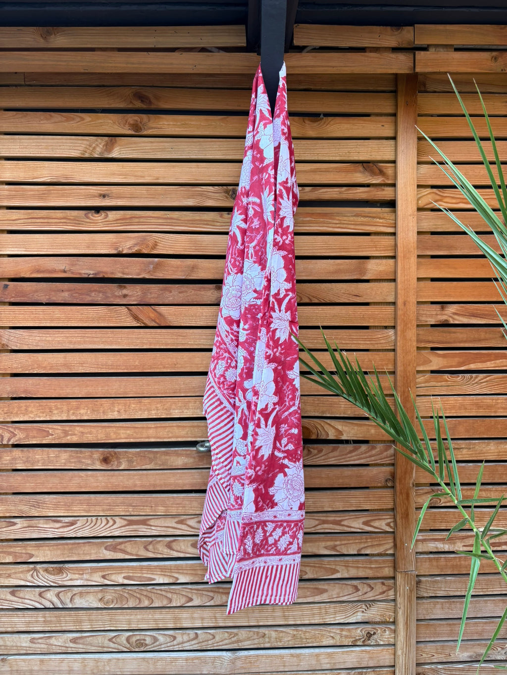Paréo en coton léger avec impression artisanale indienne de type block print. Il présente un grand motif floral détaillé aux contours blanc cassé sur un fond rouge framboise profond, structuré par de larges bordures ornées et des finitions à rayures bicolores. Le tissu est suspendu naturellement devant un mur à claire-voie en bois clair et un feuillage vert