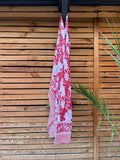Paréo en coton léger avec impression artisanale indienne de type block print. Il présente un grand motif floral détaillé aux contours blanc cassé sur un fond rouge framboise profond, structuré par de larges bordures ornées et des finitions à rayures bicolores. Le tissu est suspendu naturellement devant un mur à claire-voie en bois clair et un feuillage vert