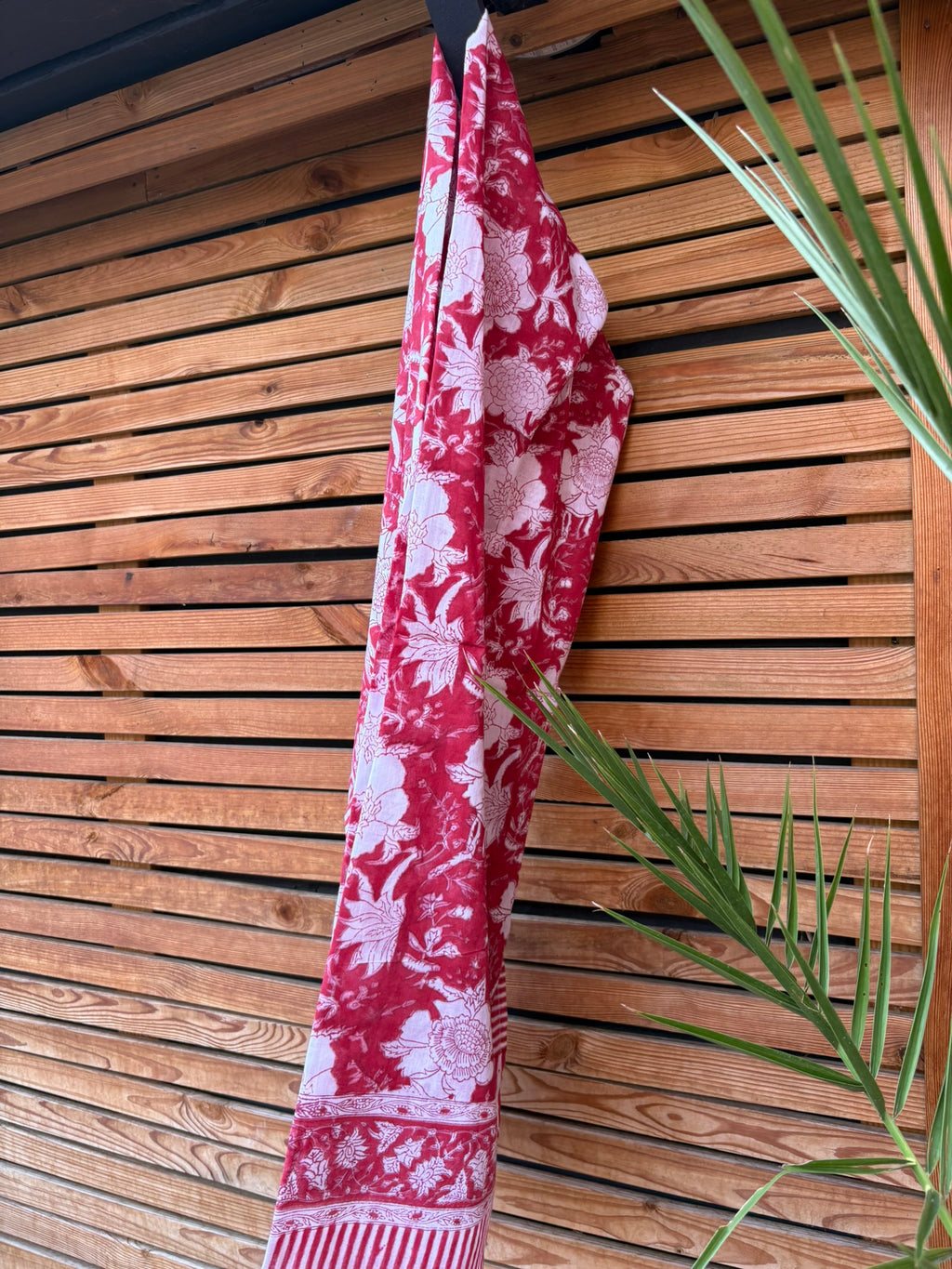 Paréo en coton léger avec impression artisanale indienne de type block print. Il présente un grand motif floral détaillé aux contours blanc cassé sur un fond rouge framboise profond, structuré par de larges bordures ornées et des finitions à rayures bicolores. Le tissu est suspendu naturellement devant un mur à claire-voie en bois clair et un feuillage vert