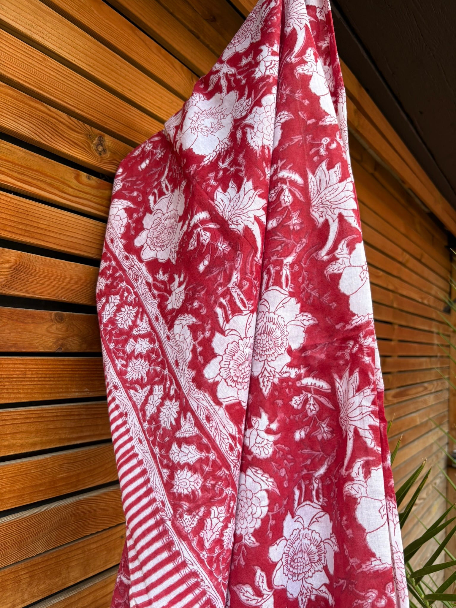 Paréo en coton léger avec impression artisanale indienne de type block print. Il présente un grand motif floral détaillé aux contours blanc cassé sur un fond rouge framboise profond, structuré par de larges bordures ornées et des finitions à rayures bicolores. Le tissu est suspendu naturellement devant un mur à claire-voie en bois clair et un feuillage vert