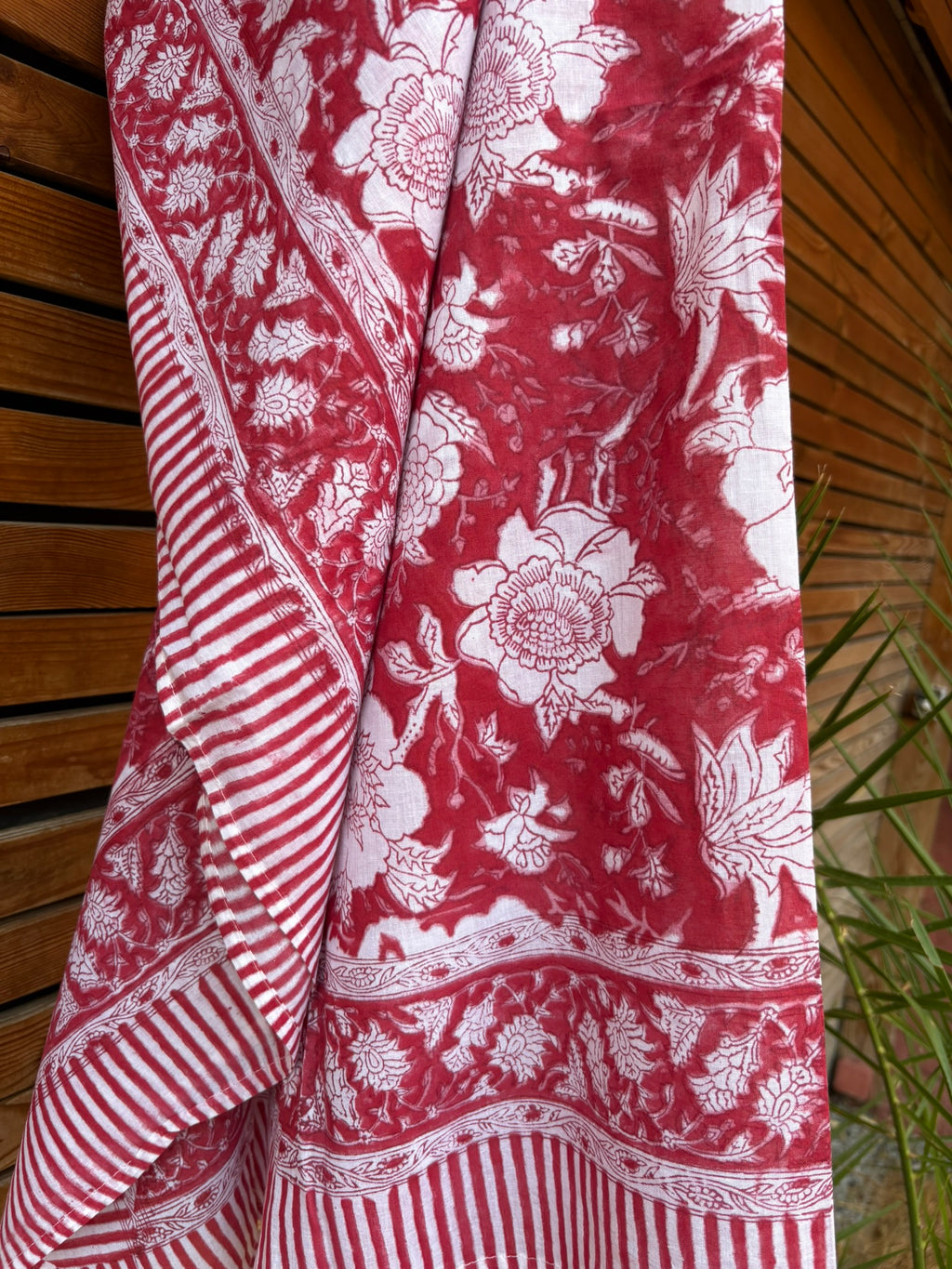 Paréo en coton léger avec impression artisanale indienne de type block print. Il présente un grand motif floral détaillé aux contours blanc cassé sur un fond rouge framboise profond, structuré par de larges bordures ornées et des finitions à rayures bicolores. Le tissu est suspendu naturellement devant un mur à claire-voie en bois clair et un feuillage vert