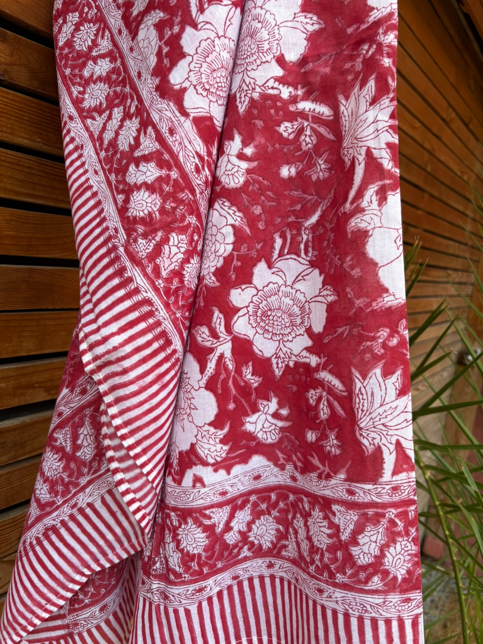 Paréo en coton léger avec impression artisanale indienne de type block print. Il présente un grand motif floral détaillé aux contours blanc cassé sur un fond rouge framboise profond, structuré par de larges bordures ornées et des finitions à rayures bicolores. Le tissu est suspendu naturellement devant un mur à claire-voie en bois clair et un feuillage vert