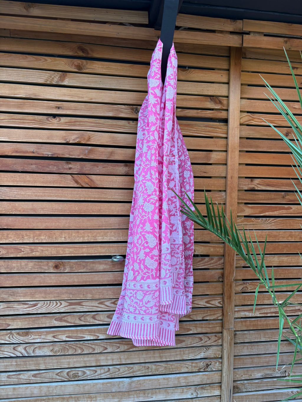 Paréo en coton léger avec impression artisanale indienne de type block print. Il présente un motif floral dense de couleur blanc sur un fond rose vif éclatant, encadré par de larges bordures ornées de motifs botaniques et de finitions à fines rayures roses et blanches. Le tissu est suspendu naturellement devant un mur à claire-voie en bois clair et un feuillage vert.
