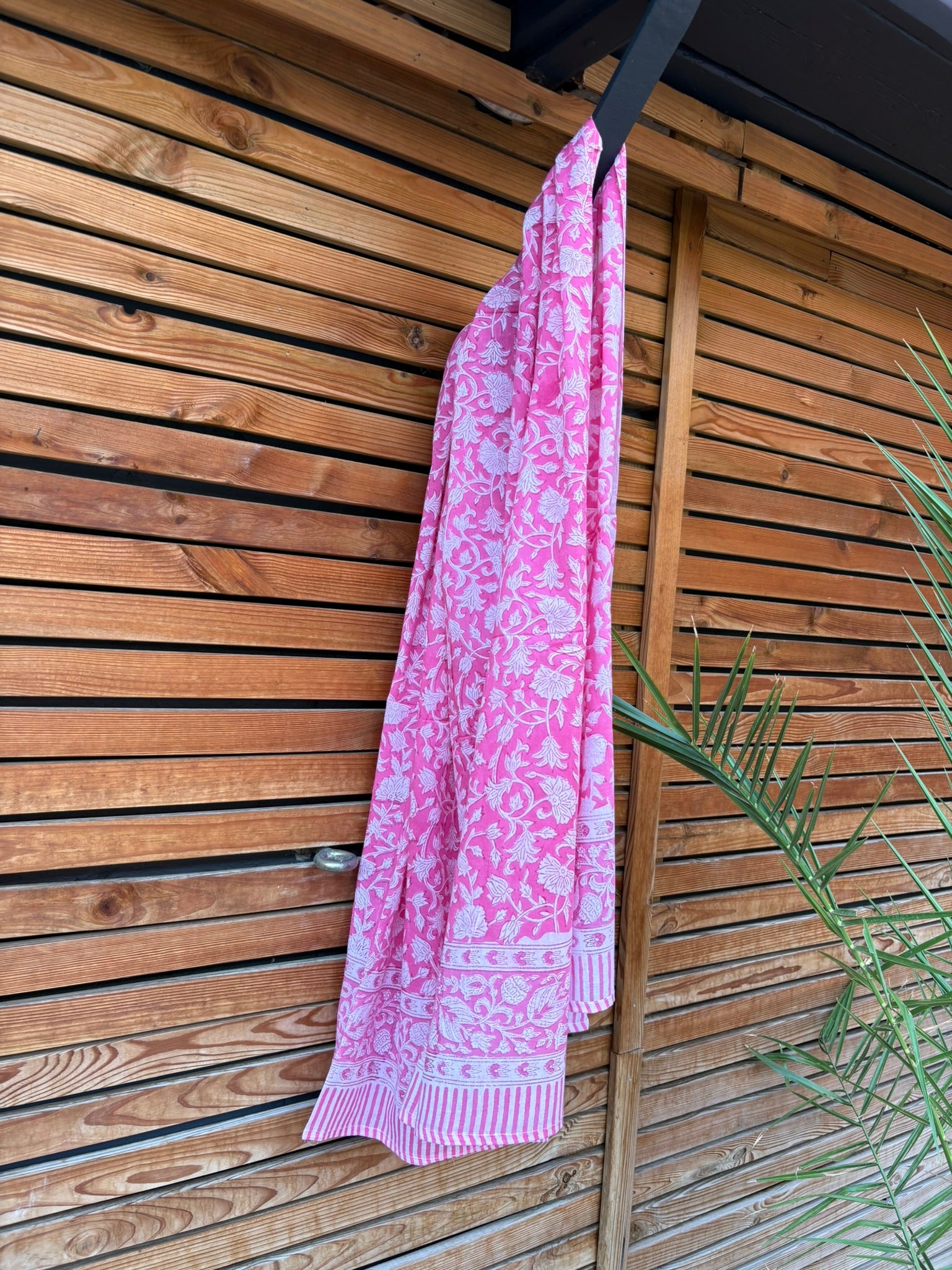 Paréo en coton léger avec impression artisanale indienne de type block print. Il présente un motif floral dense de couleur blanc sur un fond rose vif éclatant, encadré par de larges bordures ornées de motifs botaniques et de finitions à fines rayures roses et blanches. Le tissu est suspendu naturellement devant un mur à claire-voie en bois clair et un feuillage vert.