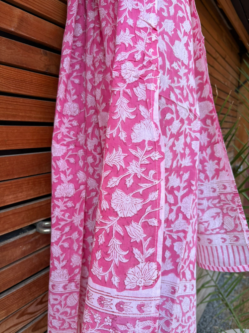 Paréo en coton léger avec impression artisanale indienne de type block print. Il présente un motif floral dense de couleur blanc sur un fond rose vif éclatant, encadré par de larges bordures ornées de motifs botaniques et de finitions à fines rayures roses et blanches. Le tissu est suspendu naturellement devant un mur à claire-voie en bois clair et un feuillage vert.