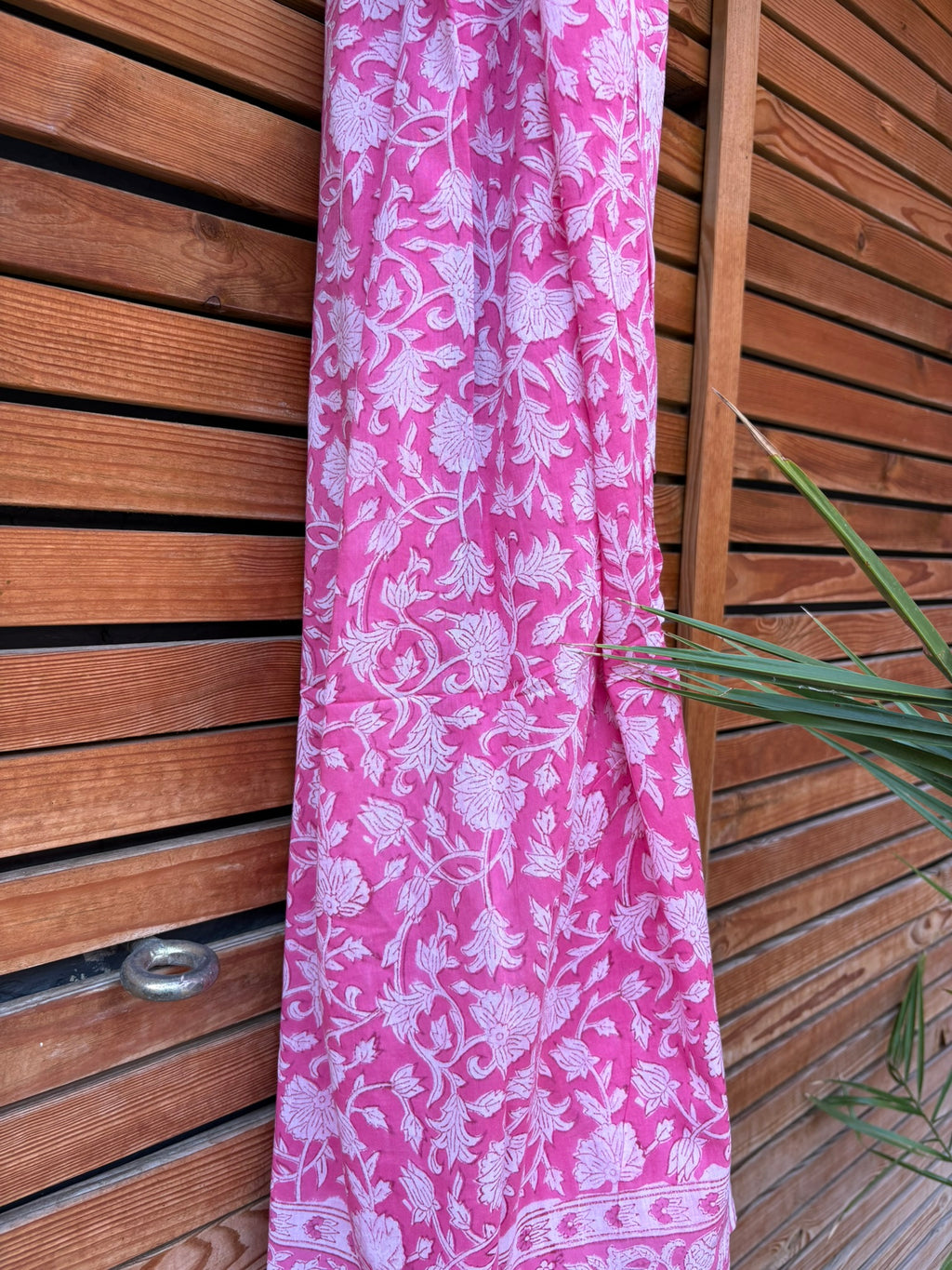 Paréo en coton léger avec impression artisanale indienne de type block print. Il présente un motif floral dense de couleur blanc sur un fond rose vif éclatant, encadré par de larges bordures ornées de motifs botaniques et de finitions à fines rayures roses et blanches. Le tissu est suspendu naturellement devant un mur à claire-voie en bois clair et un feuillage vert.
