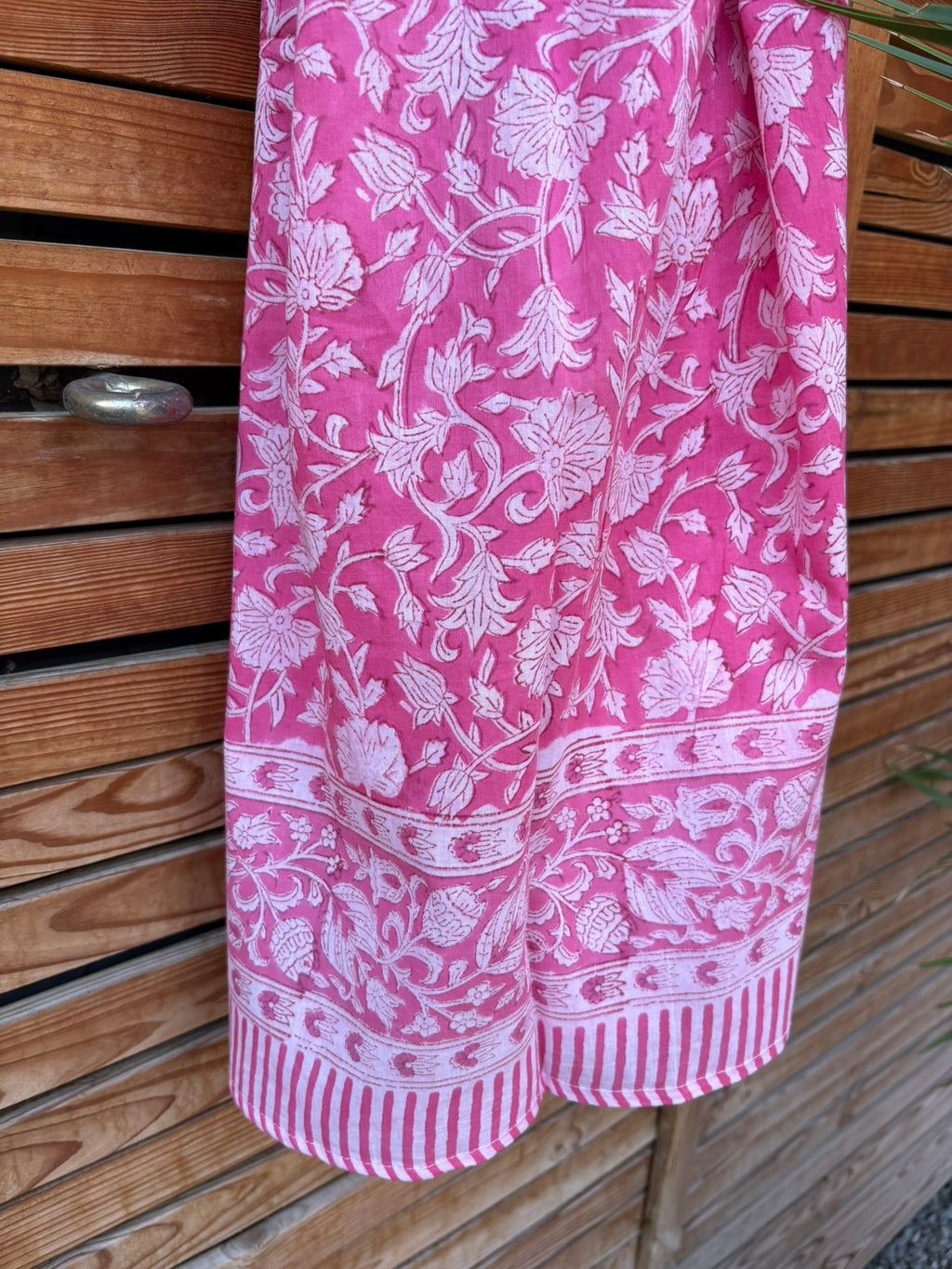 Paréo en coton léger avec impression artisanale indienne de type block print. Il présente un motif floral dense de couleur blanc sur un fond rose vif éclatant, encadré par de larges bordures ornées de motifs botaniques et de finitions à fines rayures roses et blanches. Le tissu est suspendu naturellement devant un mur à claire-voie en bois clair et un feuillage vert.