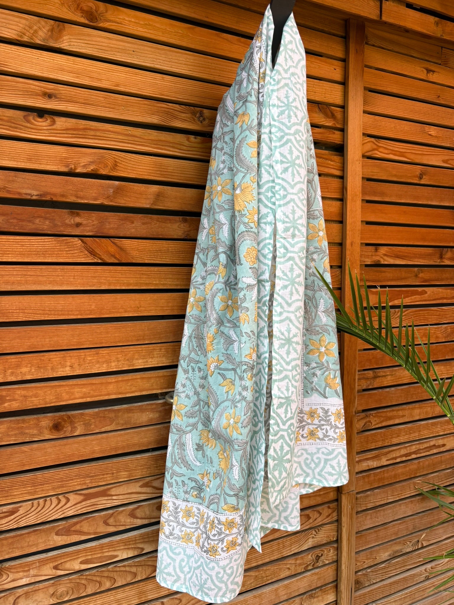 Paréo en coton léger avec impression artisanale indienne de type block print. Il présente un motif floral et végétal aux teintes jaune et gris-vert sur un fond vert d'eau clair, associé à des bandes de motifs géométriques inspirés des carreaux traditionnels. Le tissu est suspendu naturellement devant un mur à claire-voie en bois clair et un feuillage vert.