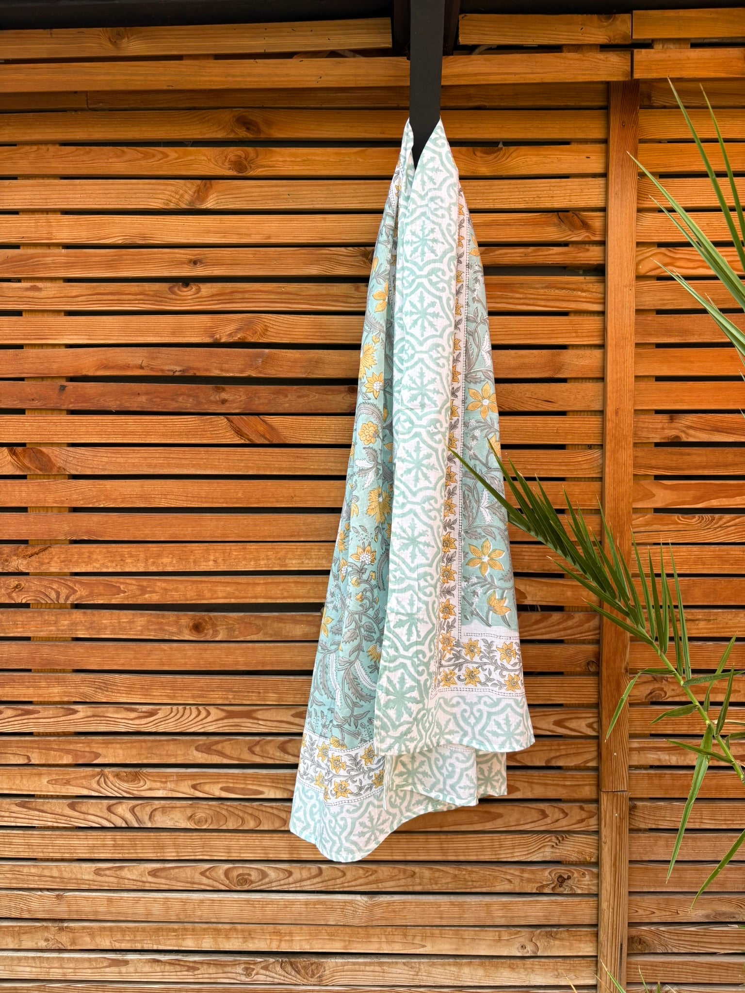 Paréo en coton léger avec impression artisanale indienne de type block print. Il présente un motif floral et végétal aux teintes jaune et gris-vert sur un fond vert d'eau clair, associé à des bandes de motifs géométriques inspirés des carreaux traditionnels. Le tissu est suspendu naturellement devant un mur à claire-voie en bois clair et un feuillage vert.