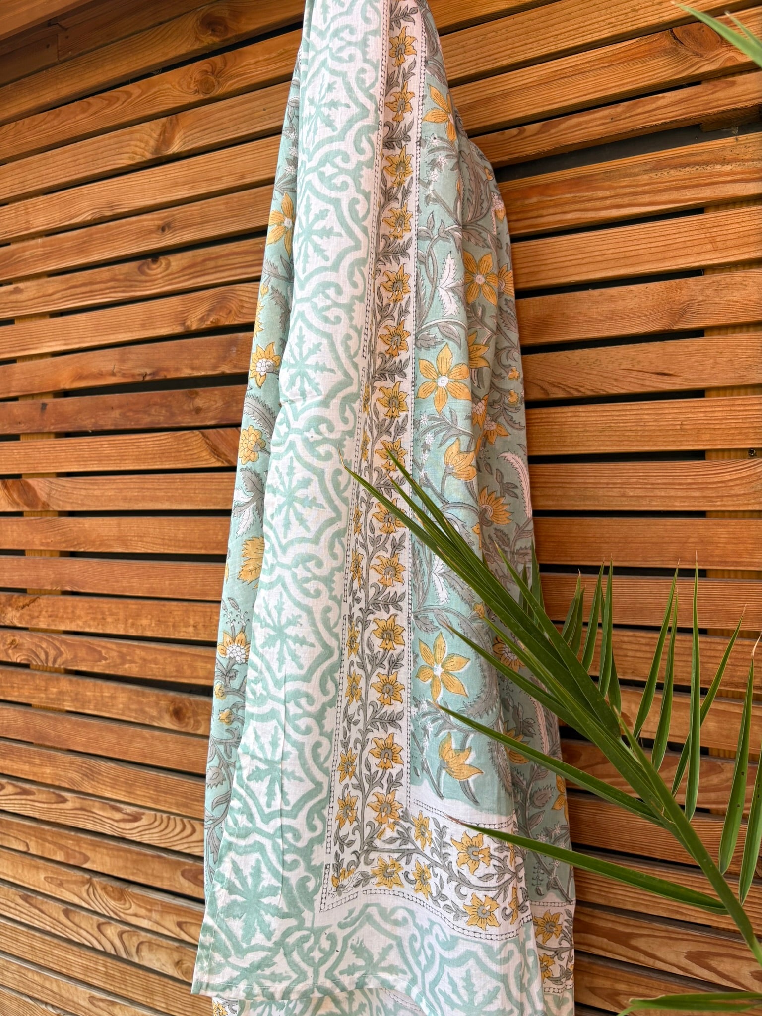 Paréo en coton léger avec impression artisanale indienne de type block print. Il présente un motif floral et végétal aux teintes jaune et gris-vert sur un fond vert d'eau clair, associé à des bandes de motifs géométriques inspirés des carreaux traditionnels. Le tissu est suspendu naturellement devant un mur à claire-voie en bois clair et un feuillage vert.