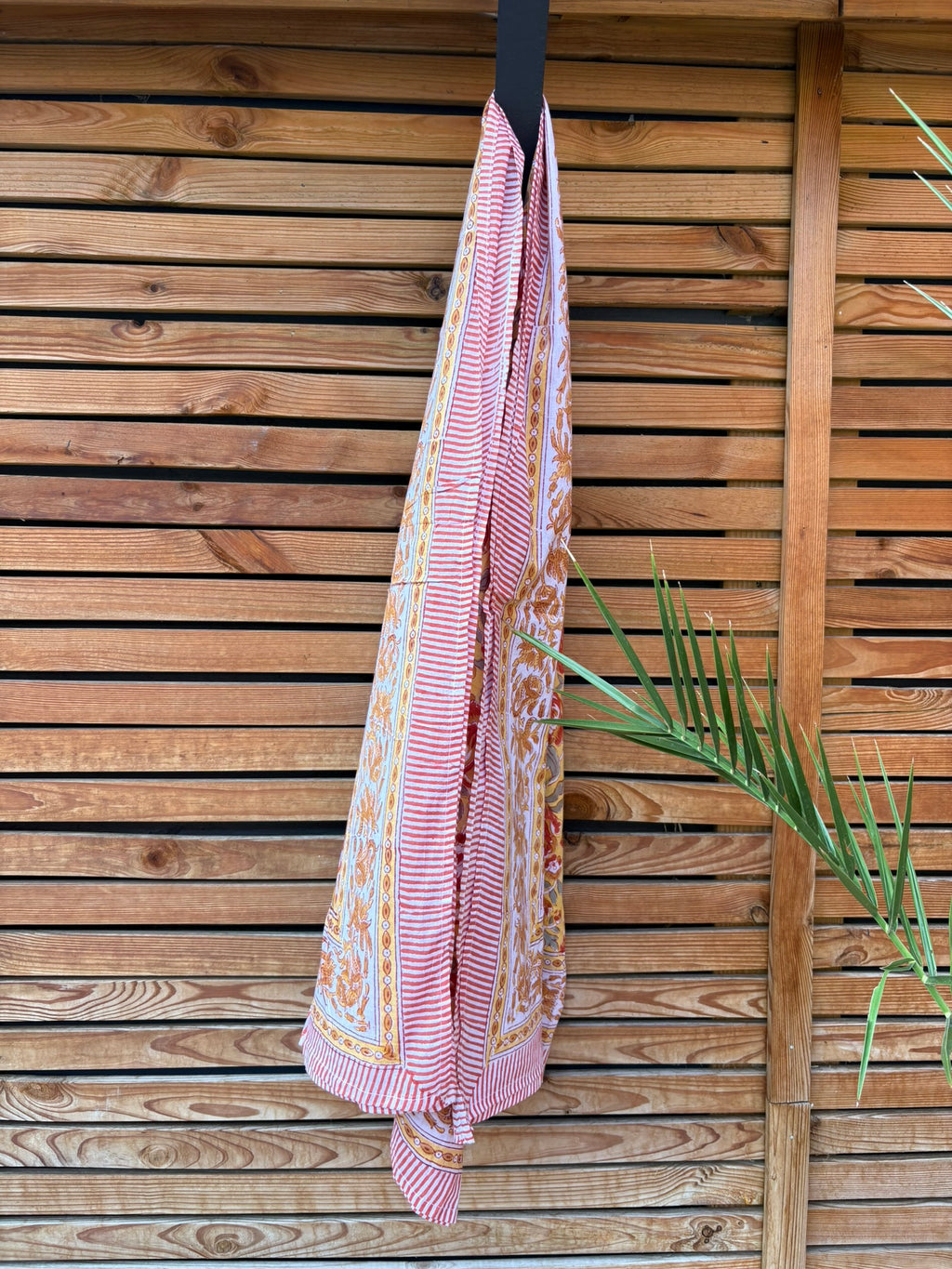 Paréo en coton léger avec impression artisanale indienne de type block print. Il présente un motif floral aux teintes chaudes jaune moutarde, orange et rouge, contrasté par de larges bordures ornementales sur fond blanc et des finitions à fines rayures rouges. Le tissu est suspendu naturellement devant un mur à claire-voie en bois clair, à côté d'un feuillage vert