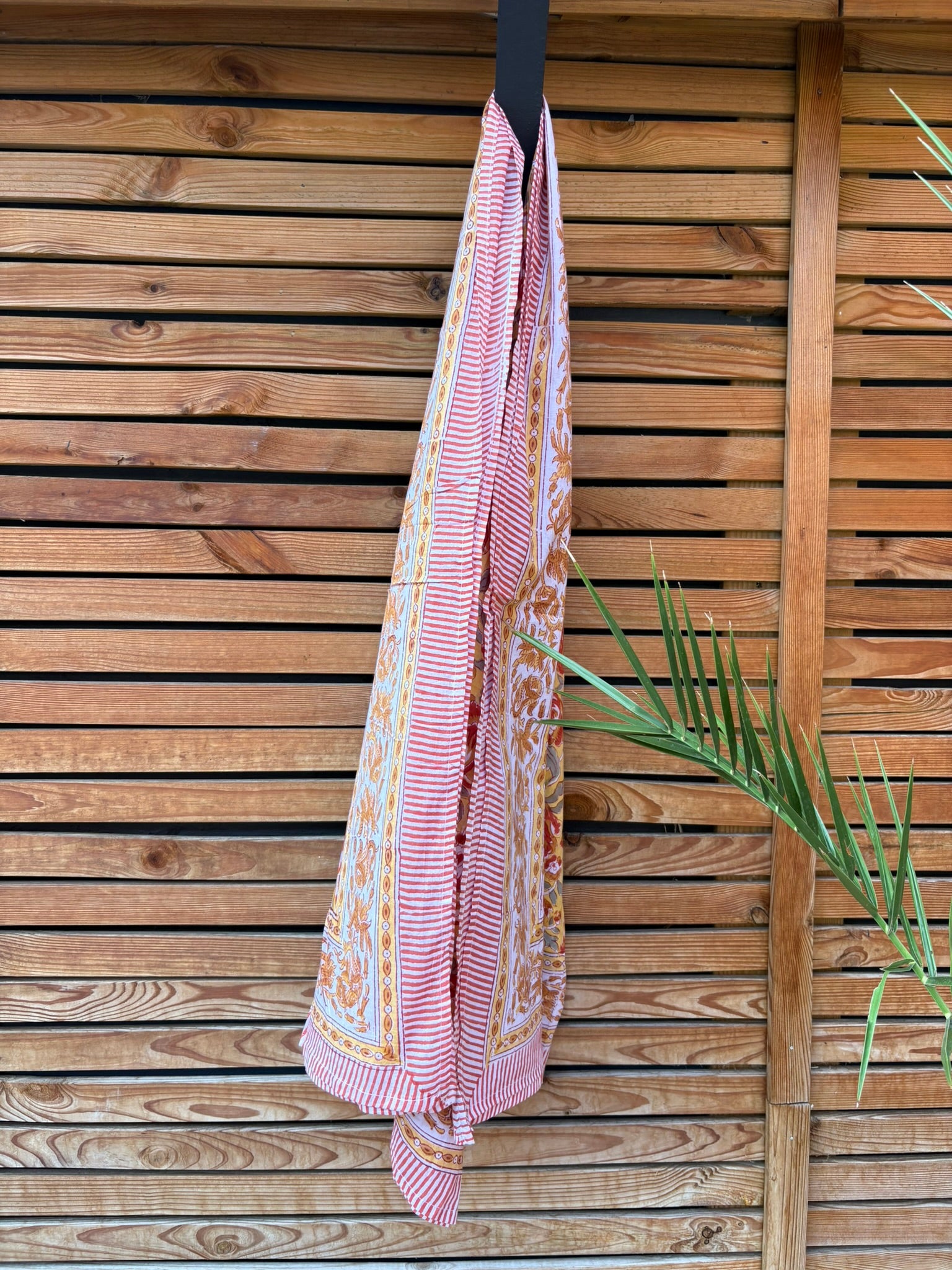 Paréo en coton léger avec impression artisanale indienne de type block print. Il présente un motif floral aux teintes chaudes jaune moutarde, orange et rouge, contrasté par de larges bordures ornementales sur fond blanc et des finitions à fines rayures rouges. Le tissu est suspendu naturellement devant un mur à claire-voie en bois clair, à côté d'un feuillage vert