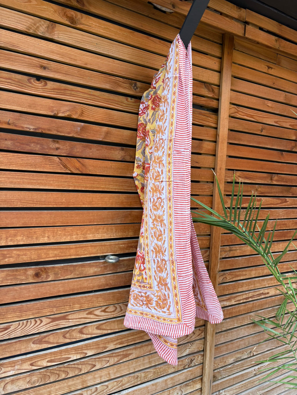 Paréo en coton léger avec impression artisanale indienne de type block print. Il présente un motif floral aux teintes chaudes jaune moutarde, orange et rouge, contrasté par de larges bordures ornementales sur fond blanc et des finitions à fines rayures rouges. Le tissu est suspendu naturellement devant un mur à claire-voie en bois clair, à côté d'un feuillage vert