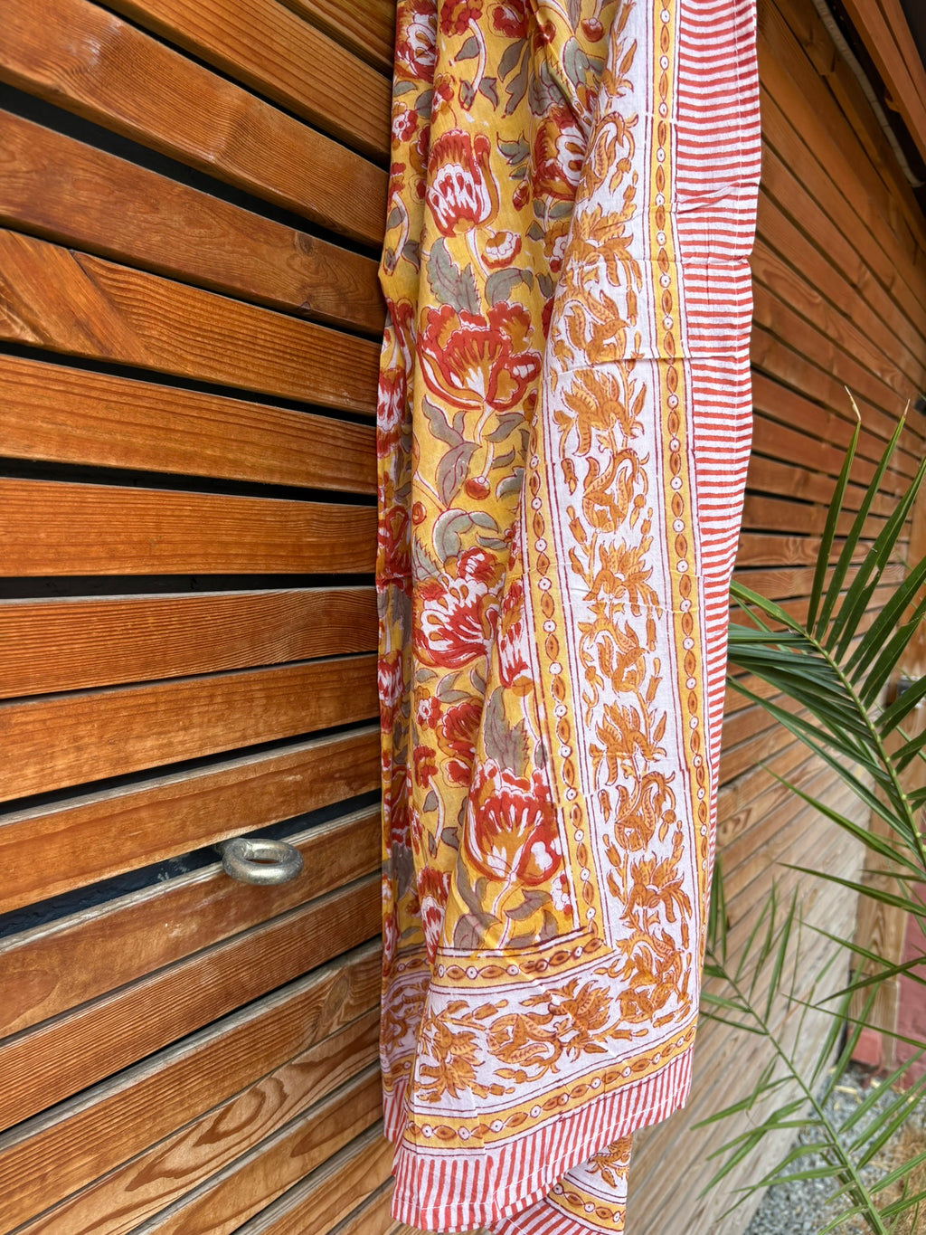 Paréo en coton léger avec impression artisanale indienne de type block print. Il présente un motif floral aux teintes chaudes jaune moutarde, orange et rouge, contrasté par de larges bordures ornementales sur fond blanc et des finitions à fines rayures rouges. Le tissu est suspendu naturellement devant un mur à claire-voie en bois clair, à côté d'un feuillage vert