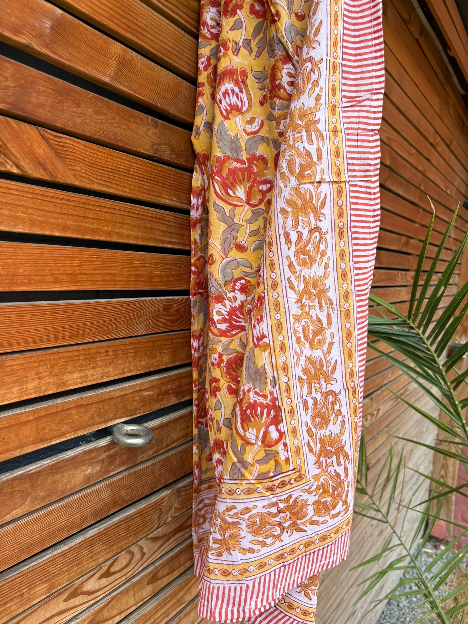 Paréo en coton léger avec impression artisanale indienne de type block print. Il présente un motif floral aux teintes chaudes jaune moutarde, orange et rouge, contrasté par de larges bordures ornementales sur fond blanc et des finitions à fines rayures rouges. Le tissu est suspendu naturellement devant un mur à claire-voie en bois clair, à côté d'un feuillage vert