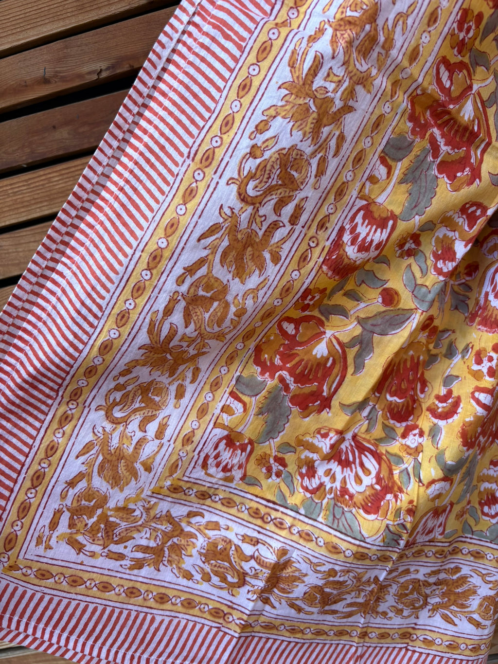 Paréo en coton léger avec impression artisanale indienne de type block print. Il présente un motif floral aux teintes chaudes jaune moutarde, orange et rouge, contrasté par de larges bordures ornementales sur fond blanc et des finitions à fines rayures rouges. 