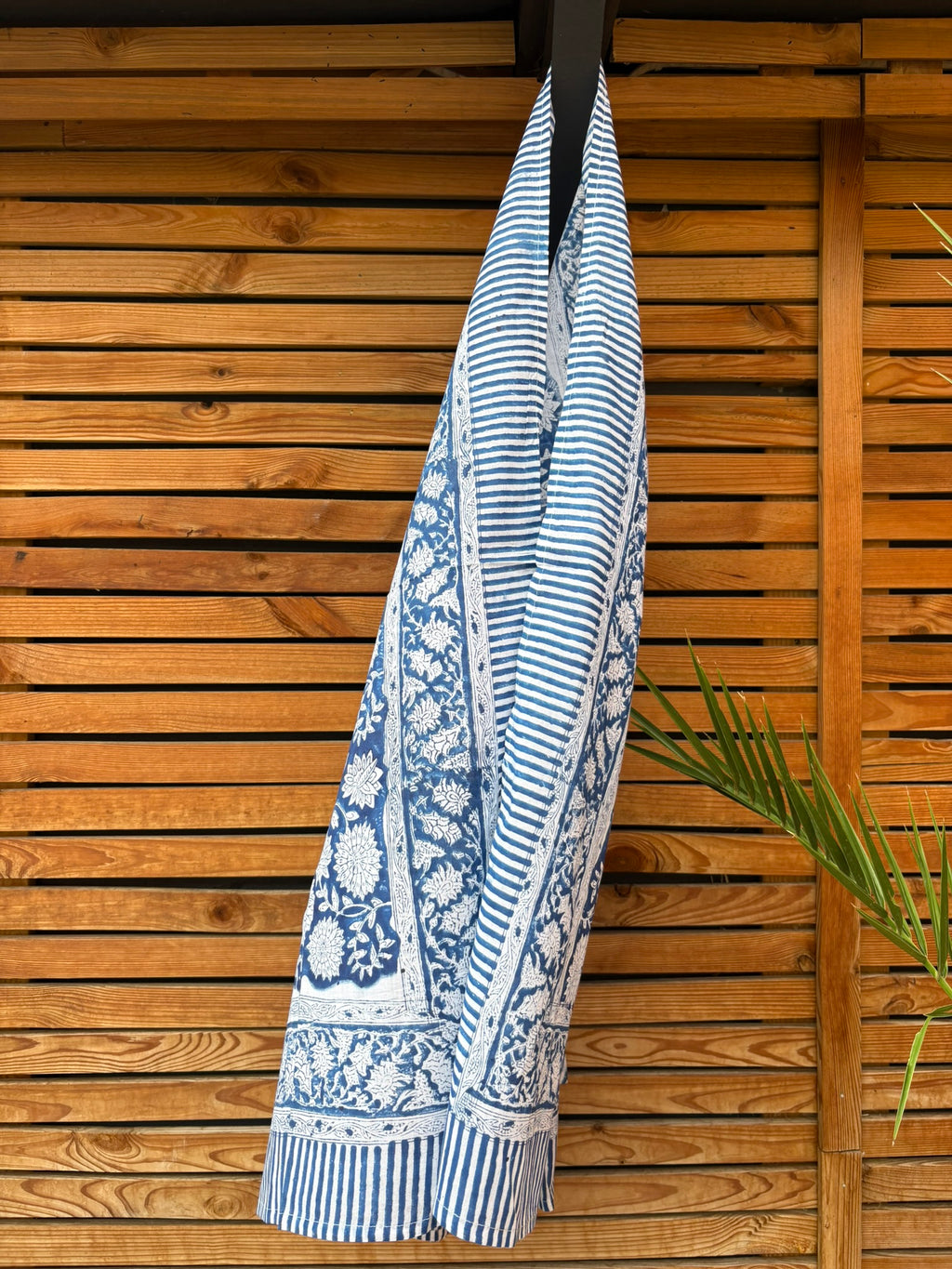 Paréo en coton léger avec impression artisanale indienne de type block print, présentant un motif floral complexe et dense de couleur blanc cassé sur un fond bleu indigo profond. Le tissu est bordé de bandes à rayures fines assorties. Il est suspendu naturellement devant un mur à claire-voie en bois clair et un feuillage vert.