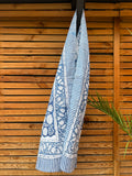 Paréo en coton léger avec impression artisanale indienne de type block print, présentant un motif floral complexe et dense de couleur blanc cassé sur un fond bleu indigo profond. Le tissu est bordé de bandes à rayures fines assorties. Il est suspendu naturellement devant un mur à claire-voie en bois clair et un feuillage vert.