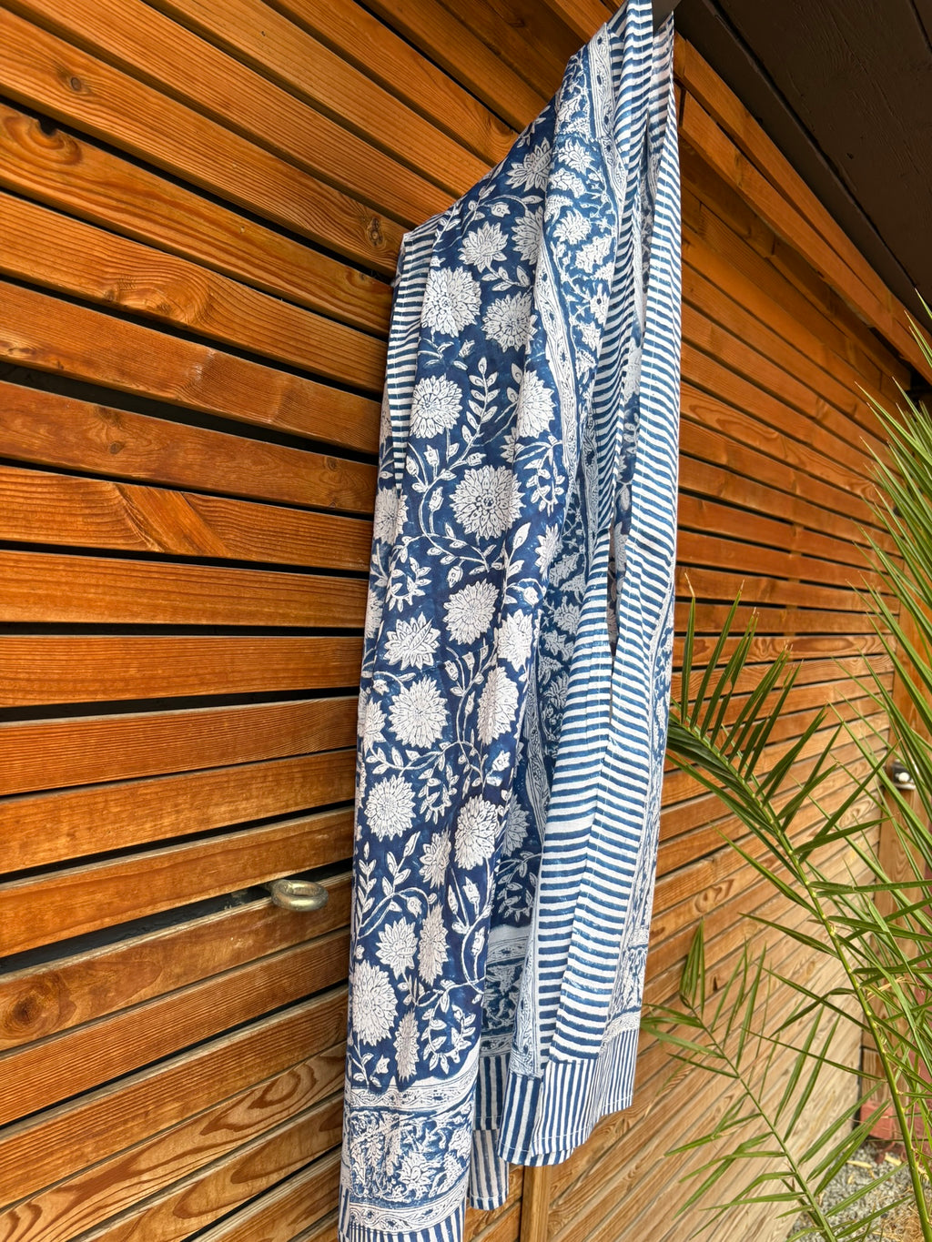 Paréo en coton léger avec impression artisanale indienne de type block print, présentant un motif floral complexe et dense de couleur blanc cassé sur un fond bleu indigo profond. Le tissu est bordé de bandes à rayures fines assorties. Il est suspendu naturellement devant un mur à claire-voie en bois clair et un feuillage vert.