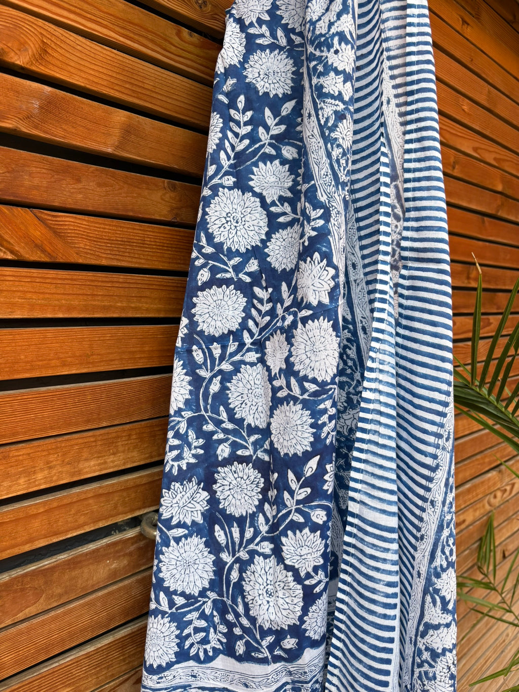 Paréo en coton léger avec impression artisanale indienne de type block print, présentant un motif floral complexe et dense de couleur blanc cassé sur un fond bleu indigo profond. Le tissu est bordé de bandes à rayures fines assorties. Il est suspendu naturellement devant un mur à claire-voie en bois clair et un feuillage vert.