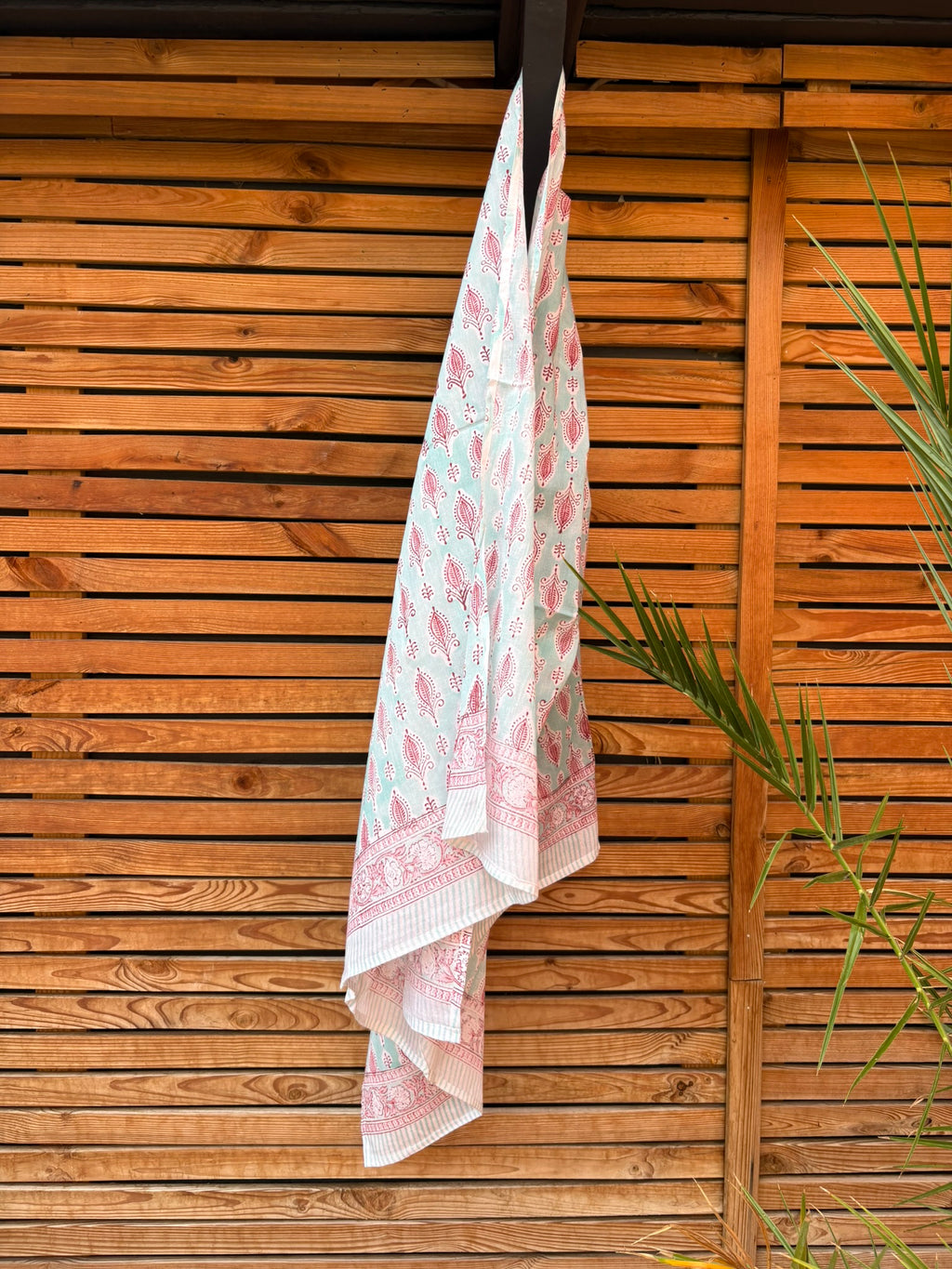 Paréo en coton léger avec impression artisanale indienne de type block print, présentant un motif répétitif de petits motifs floraux/botaniques de couleur rose fuchsia sur un fond vert menthe très clair. Une fine bordure ornementale assortie termine le tissu. Le paréo est suspendu naturellement devant un mur à claire-voie en bois clair et un feuillage vert.