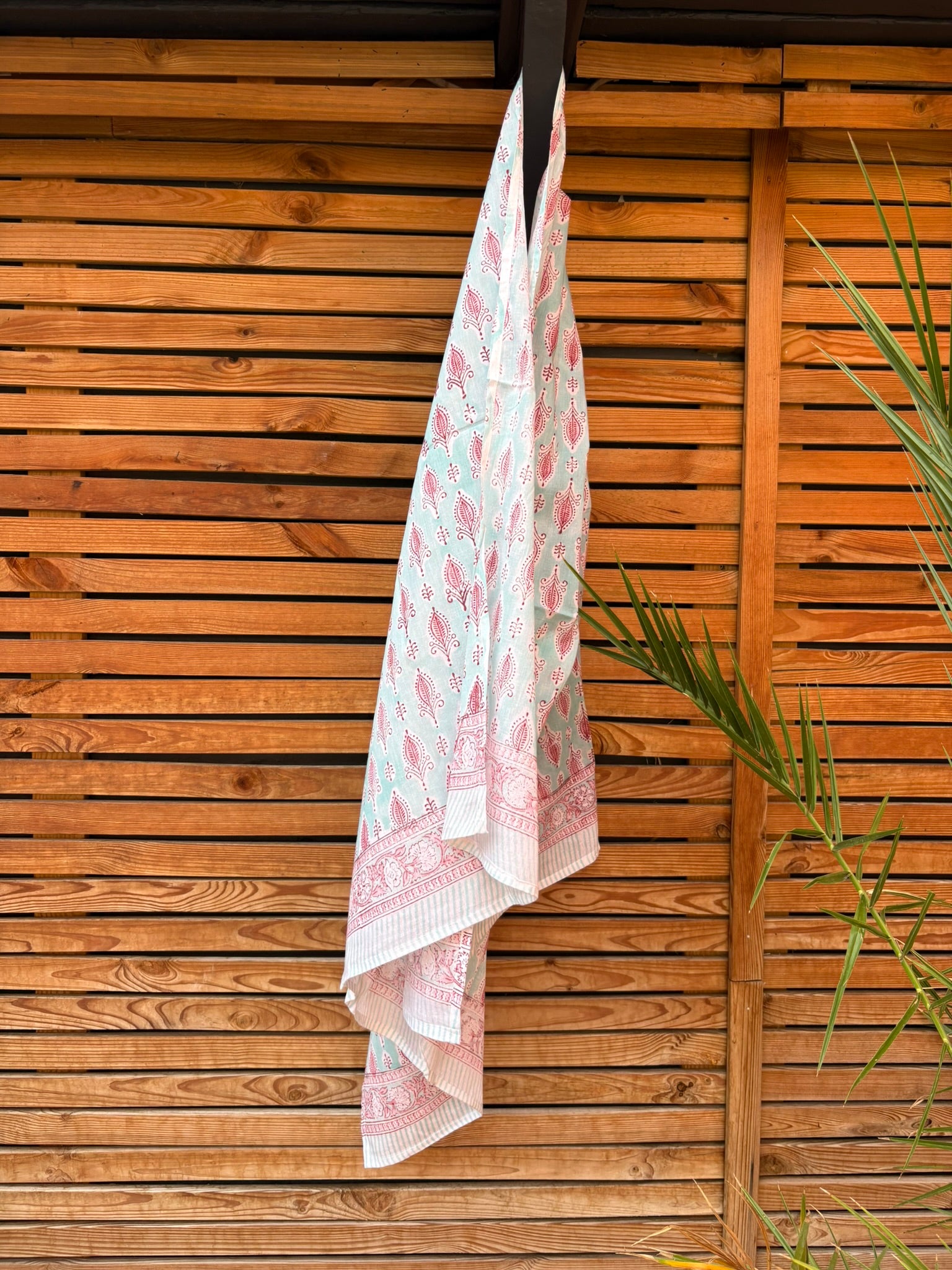 Paréo en coton léger avec impression artisanale indienne de type block print, présentant un motif répétitif de petits motifs floraux/botaniques de couleur rose fuchsia sur un fond vert menthe très clair. Une fine bordure ornementale assortie termine le tissu. Le paréo est suspendu naturellement devant un mur à claire-voie en bois clair et un feuillage vert.